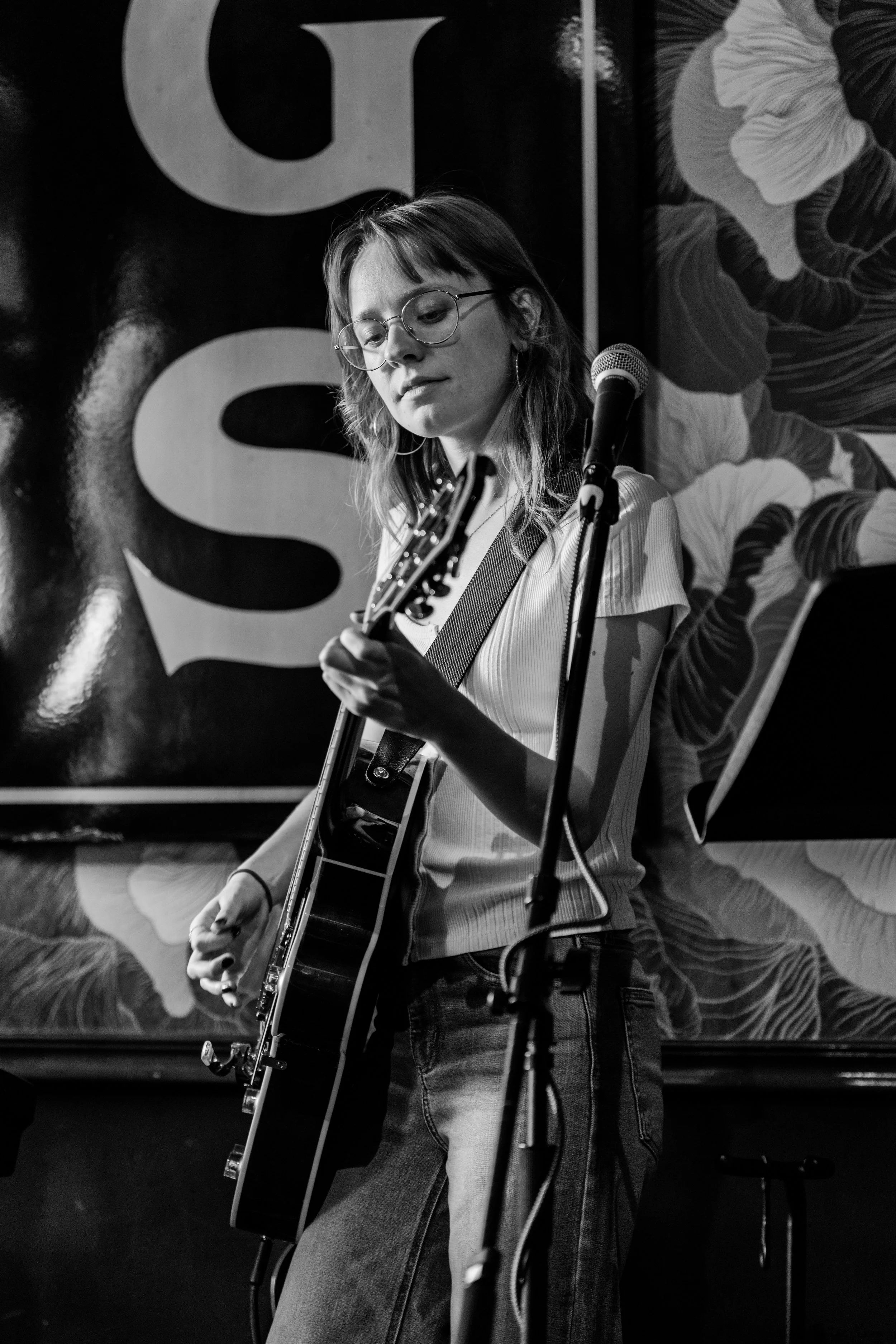 Black and white photo of a young woman with glasses playing an acoustic guitar on stage with a microphone in front of her.