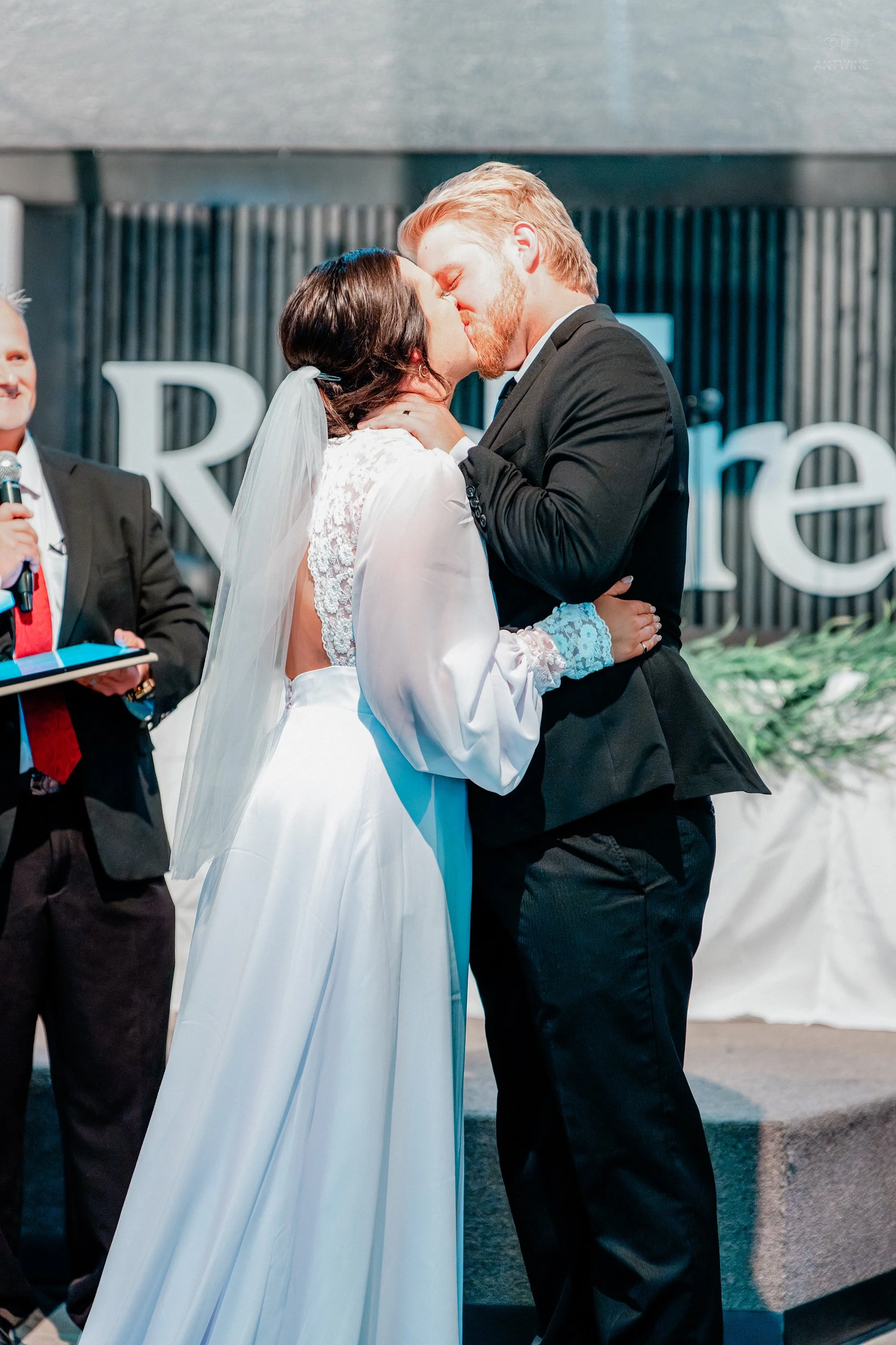 A bride and groom kissing during their wedding ceremony, with a man holding a microphone on the left and a blurred background with decorative elements.