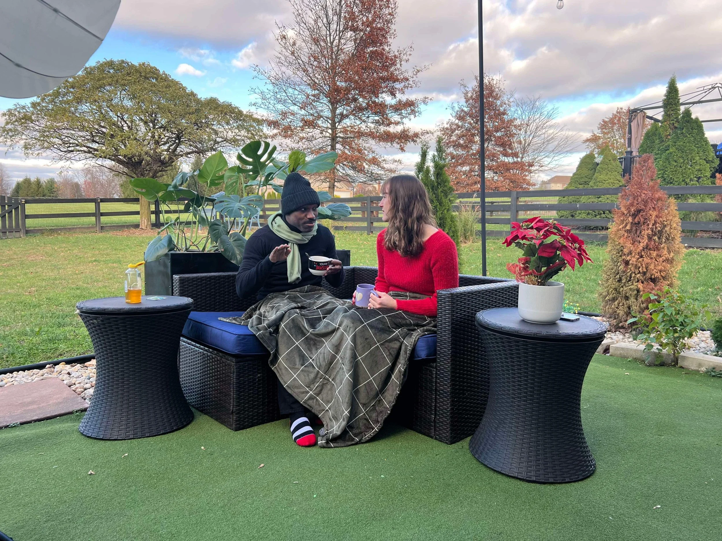 Two people sitting on a black outdoor sofa having a conversation, with two side tables, in a backyard with trees and fencing, some with autumn foliage, during a cloudy day.