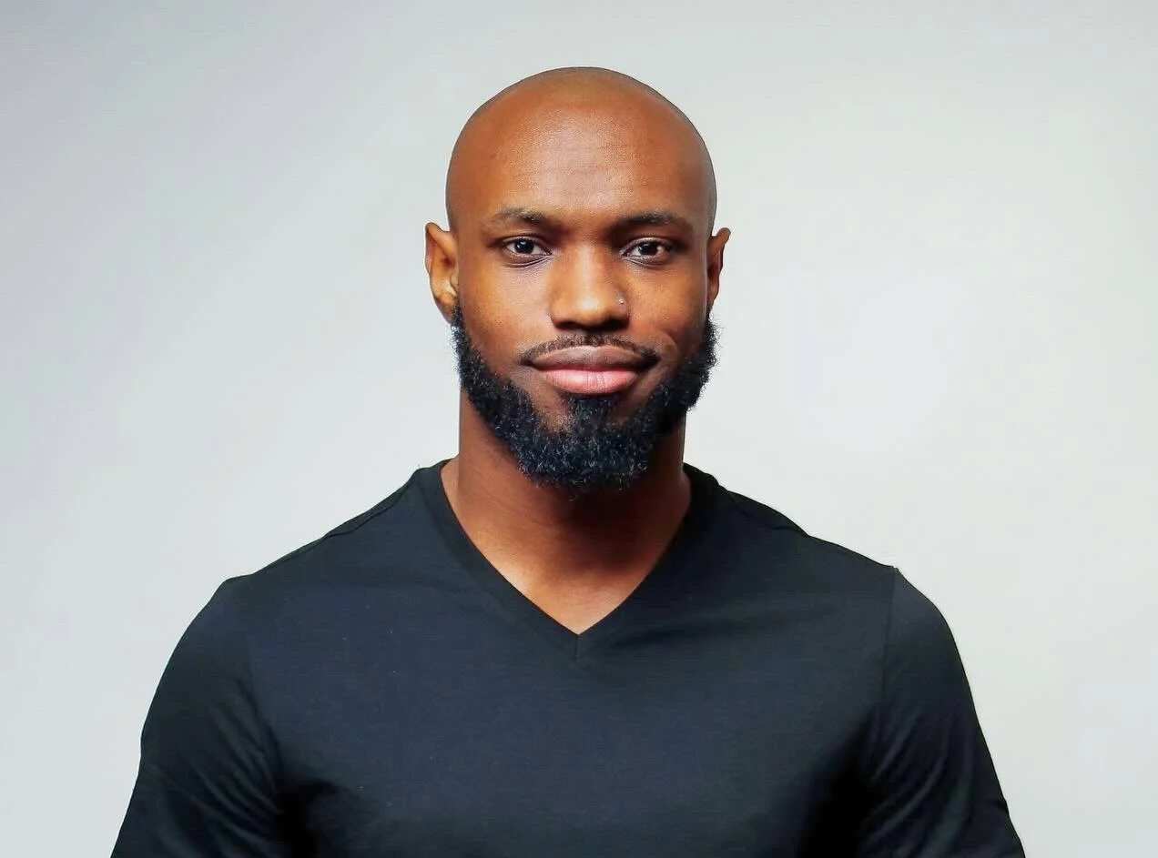 A smiling man with a shaved head, beard, and mustache, wearing a black V-neck shirt, standing against a plain light gray background.