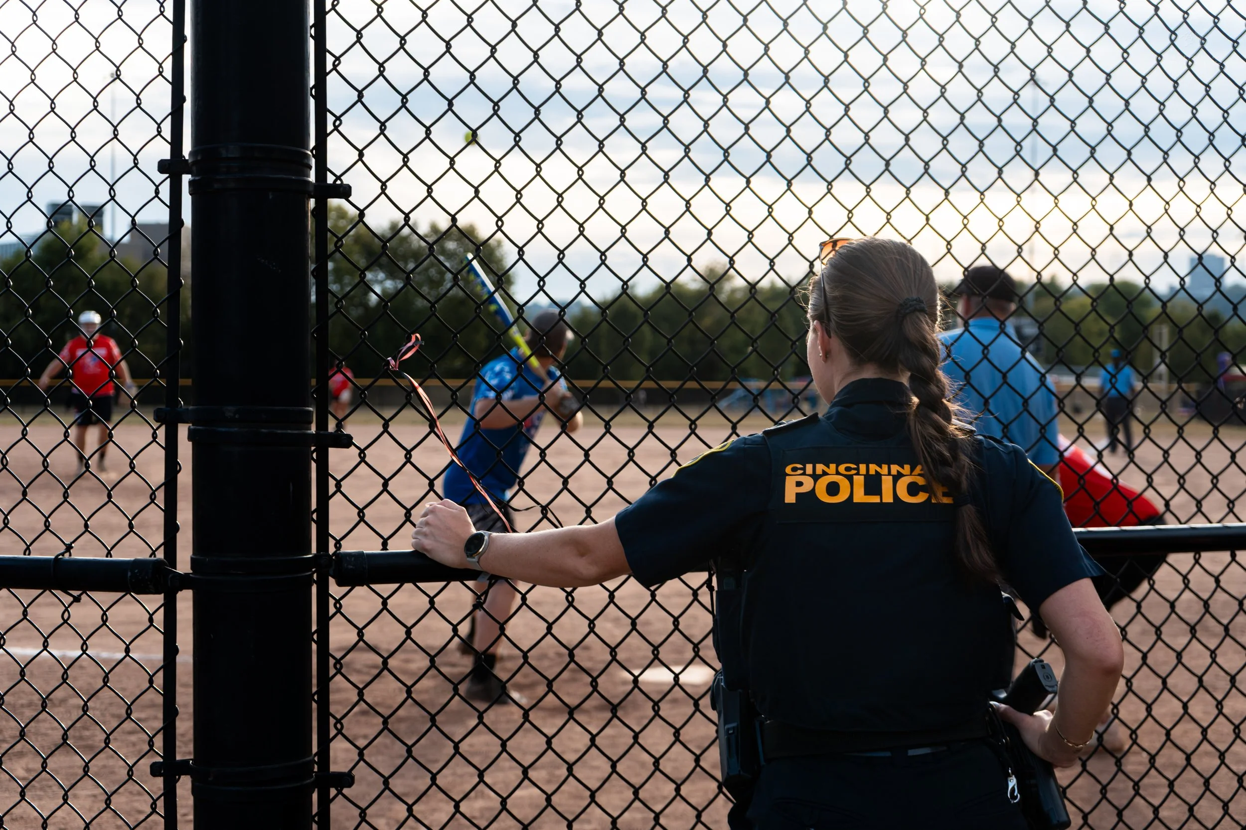 Cincinnati police officer watching children play baseball through a chain-link fence on a sports field during sunset.