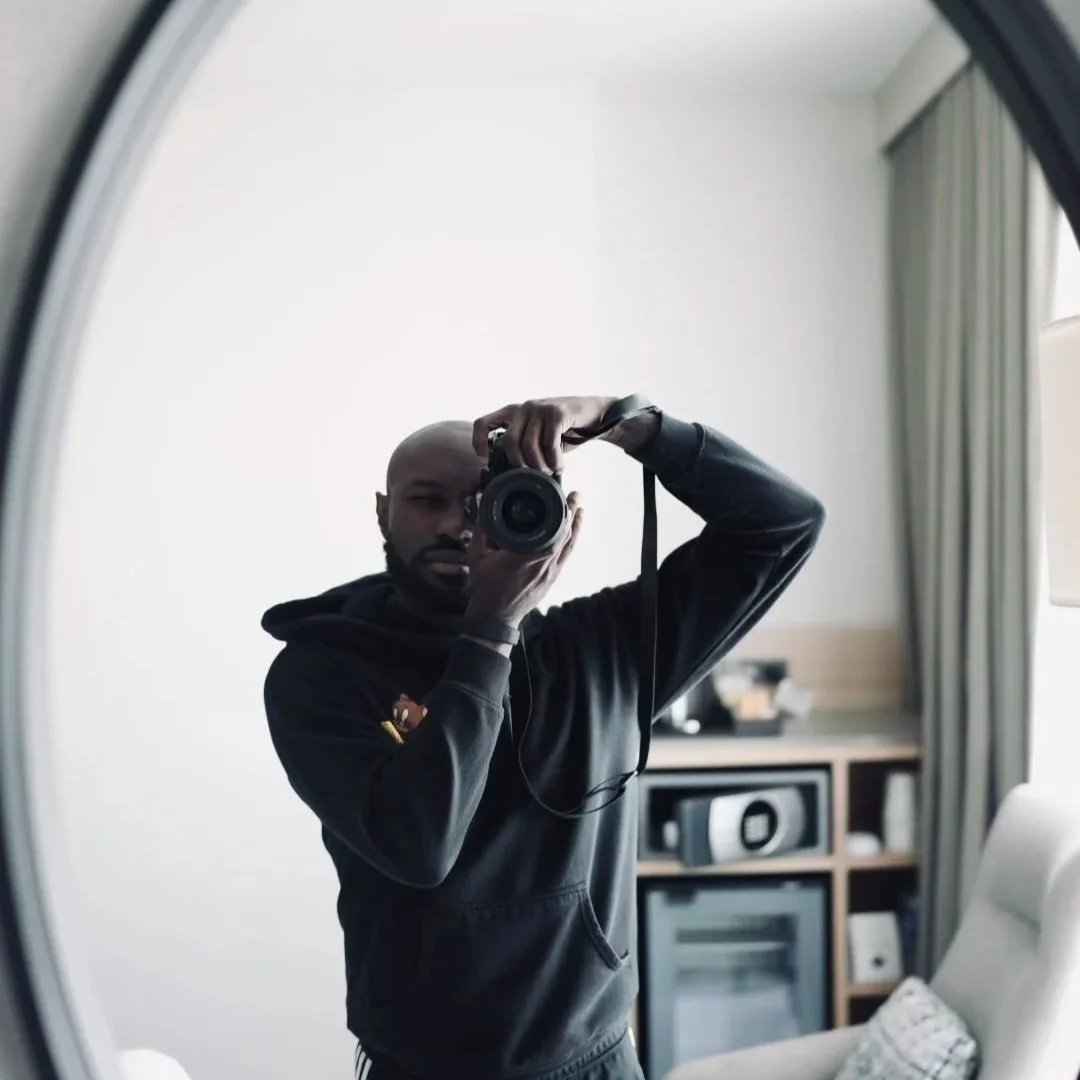A person taking a photo of themselves in a mirror, with a camera in hand. The person is wearing a hoodie, and the background shows a room with a television, a bookshelf or cabinet, and a chair.