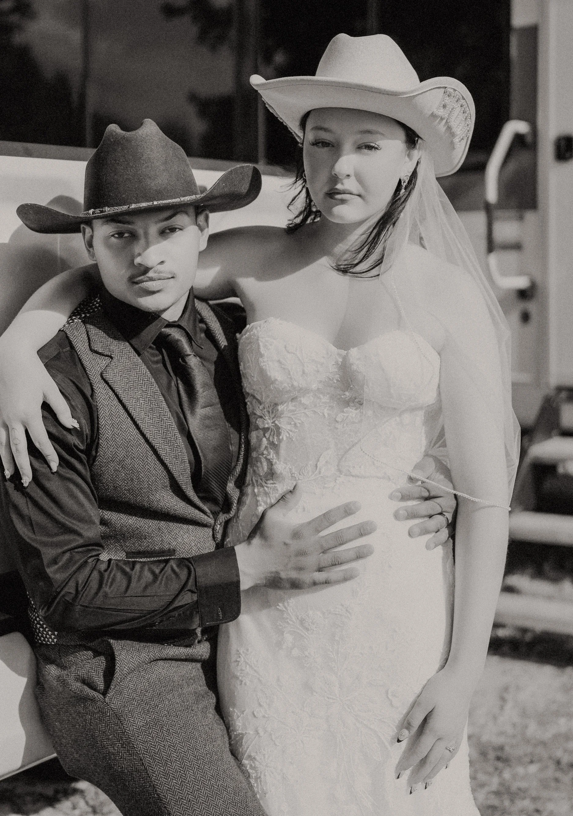 A man in western attire and a woman in a wedding dress with cowboy hats pose together outdoors.