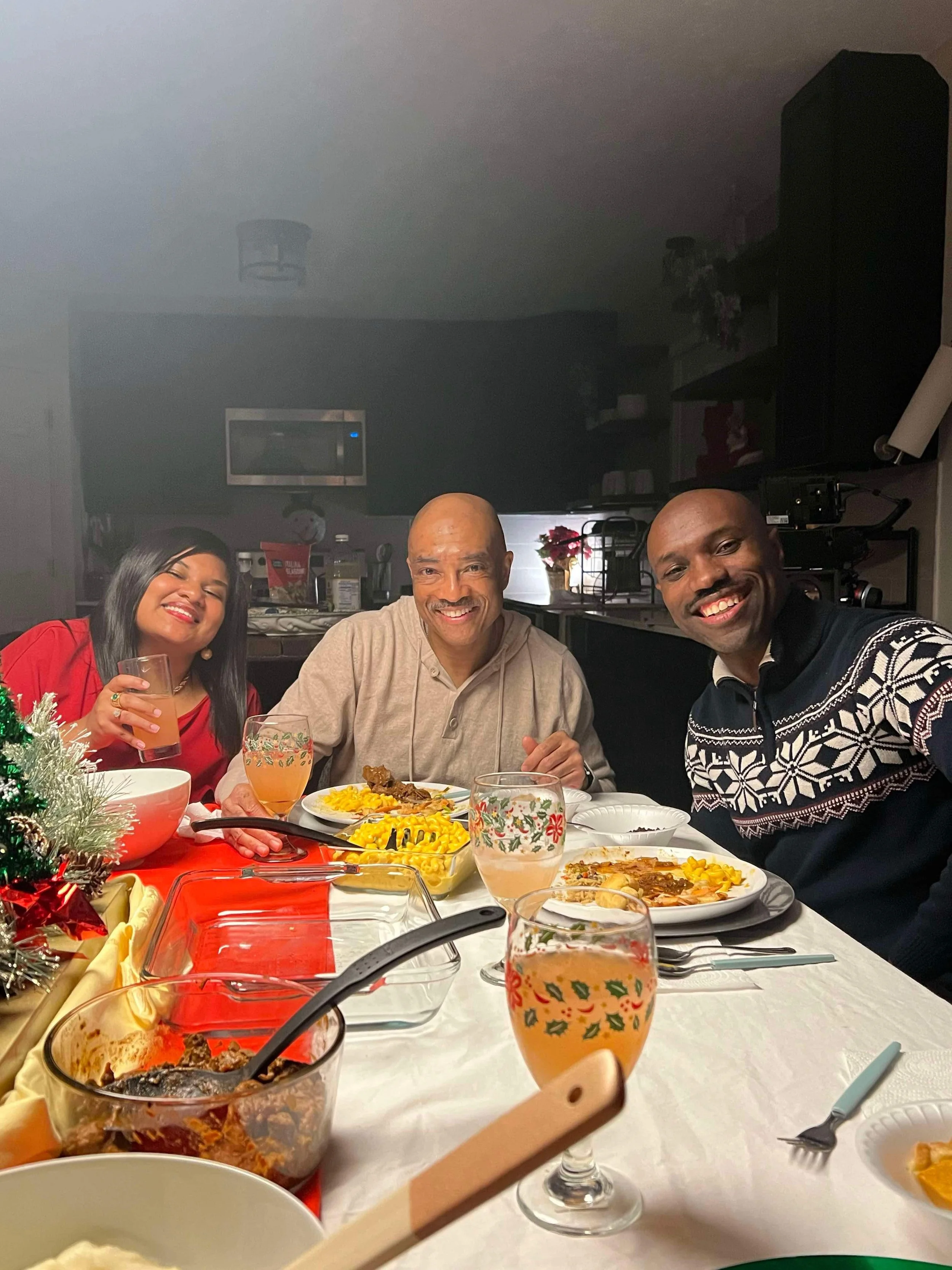 A family gathered around a dinner table celebrating Christmas. There are three adults smiling at the camera, with plates of food including yellow rice and meat. The table has drinks, a small decorated Christmas tree, and holiday-themed glassware.