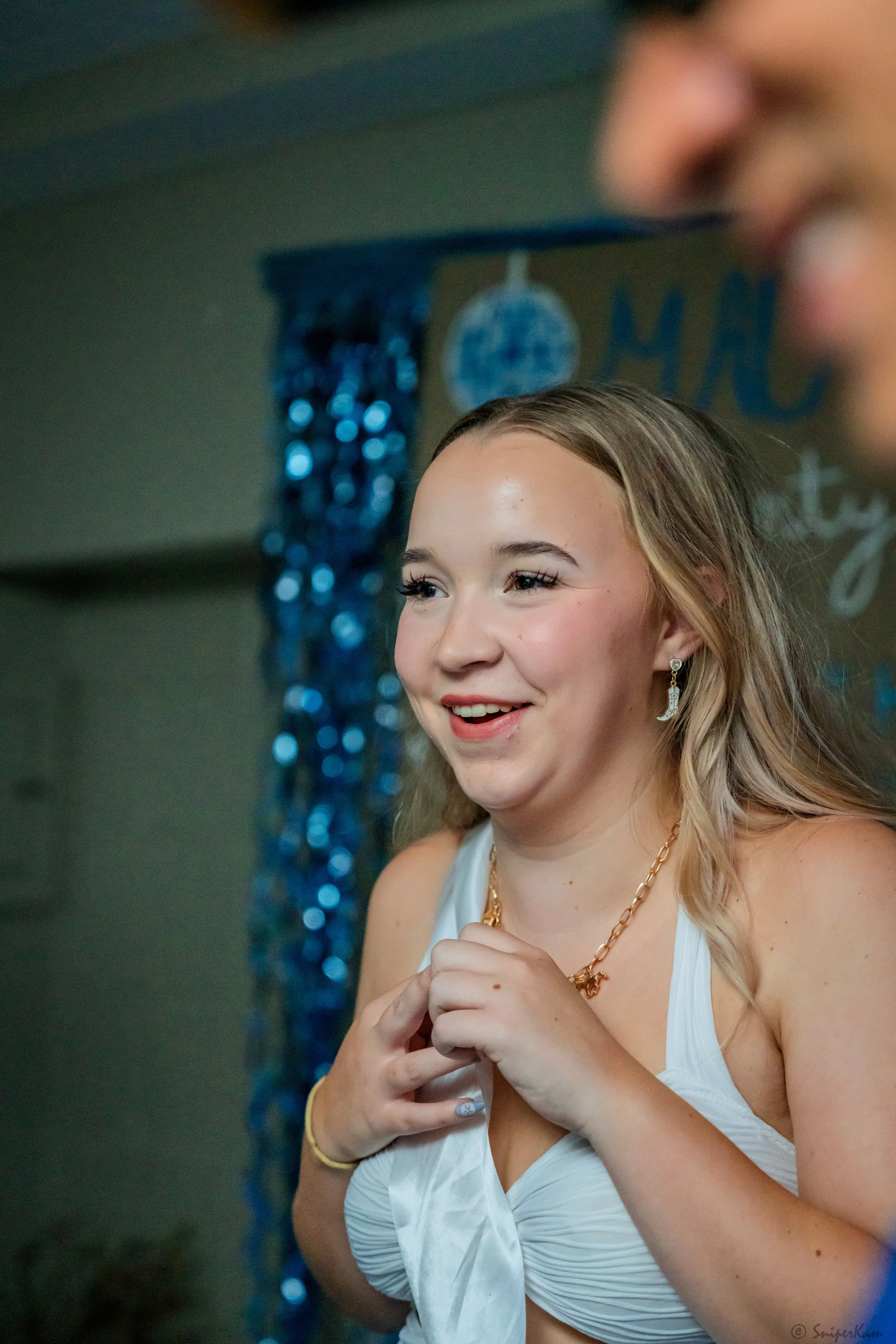 A young woman with blonde hair and light skin, smiling and wearing a white top, gold jewelry, and earrings, at a celebration or party with blue decorations in the background.
