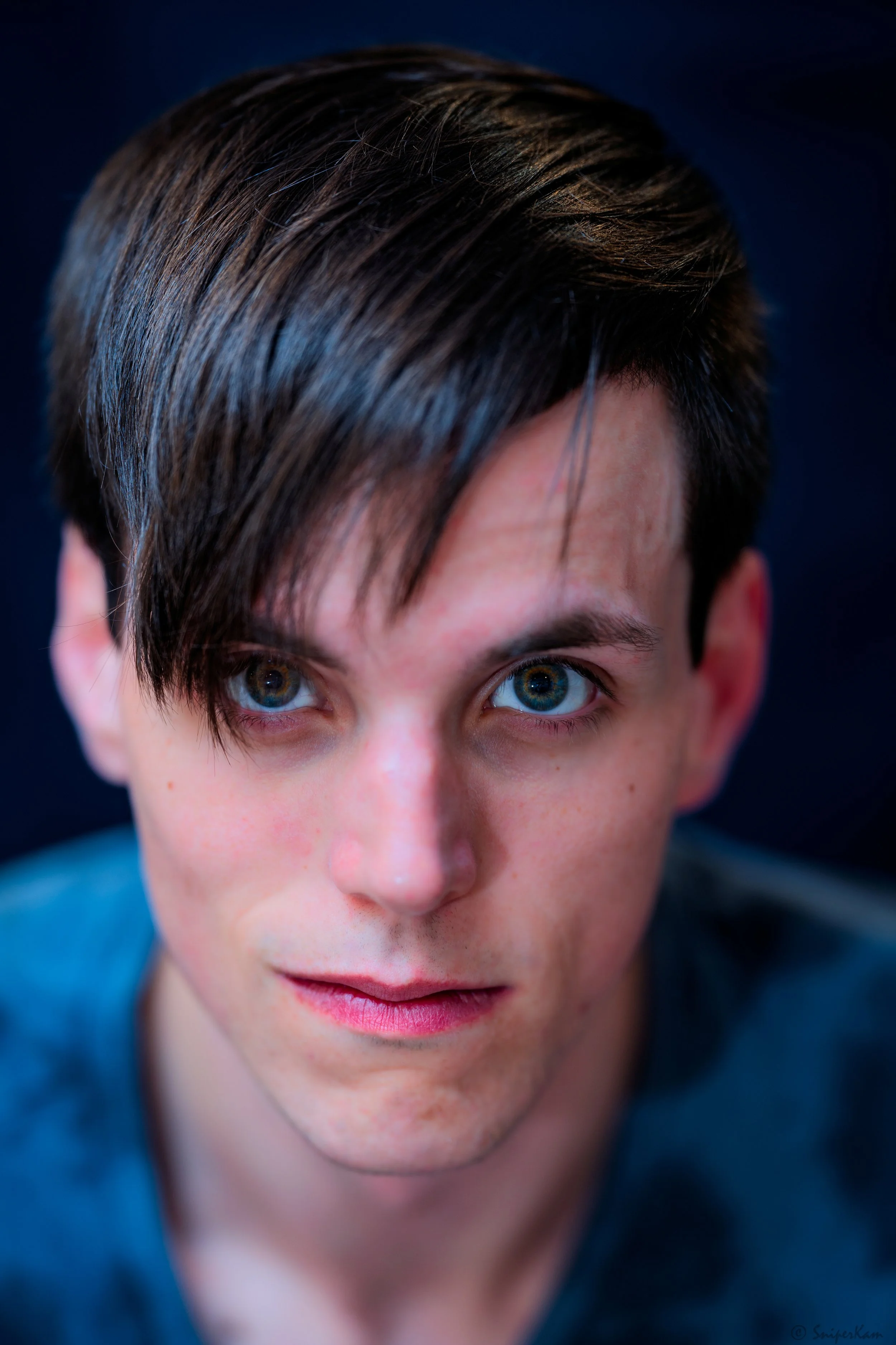 Close-up of a young man with dark hair and blue eyes, looking directly at the camera with a serious expression.