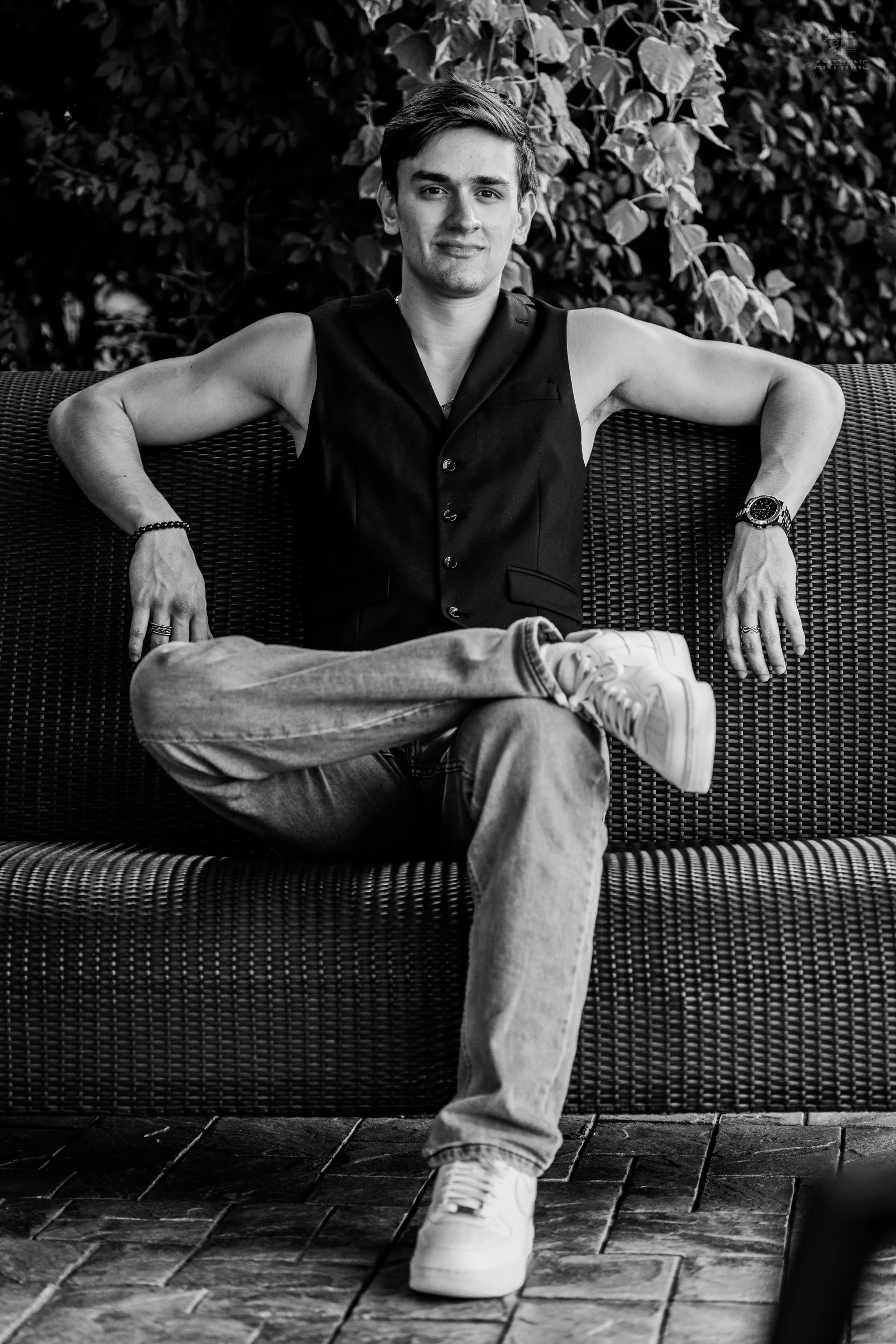 A young man with short hair sitting on a wicker outdoor sofa with leafy plants in the background. He is wearing a sleeveless black vest, jeans, white sneakers, and a wristwatch, with his arms resting on the back of the sofa.
