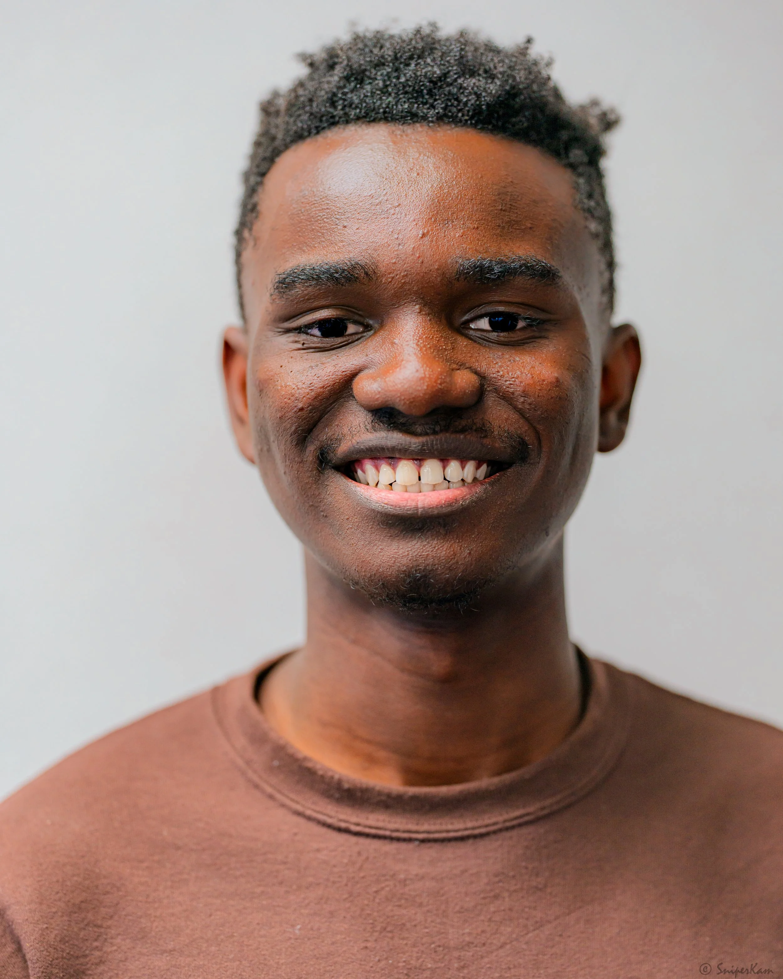 Close-up photo of a young African American man smiling with his teeth showing, wearing a brown t-shirt, against a plain light gray background.