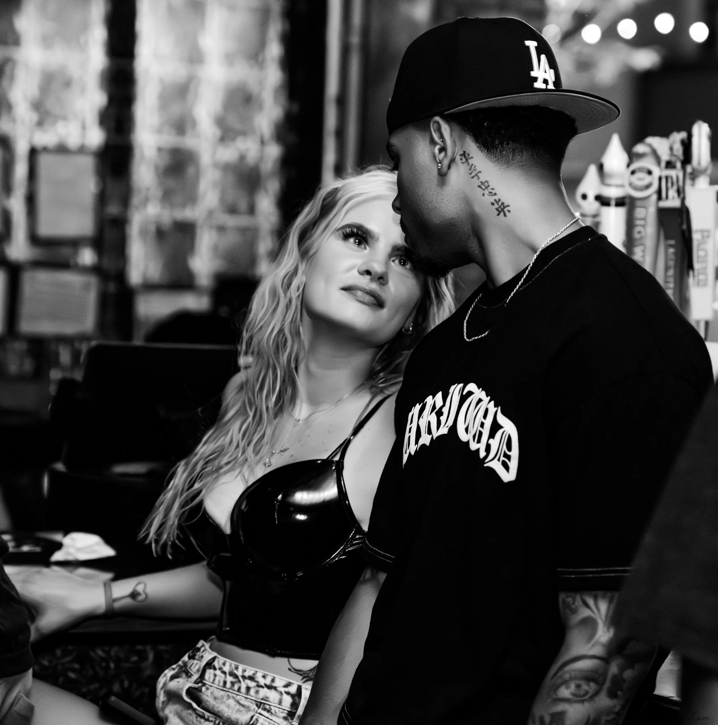A black-and-white photo of a woman with curly blonde hair looking up at a man wearing a baseball cap and T-shirt, who is leaning in close to her. The background shows a brick wall and some bottles or objects on a shelf.