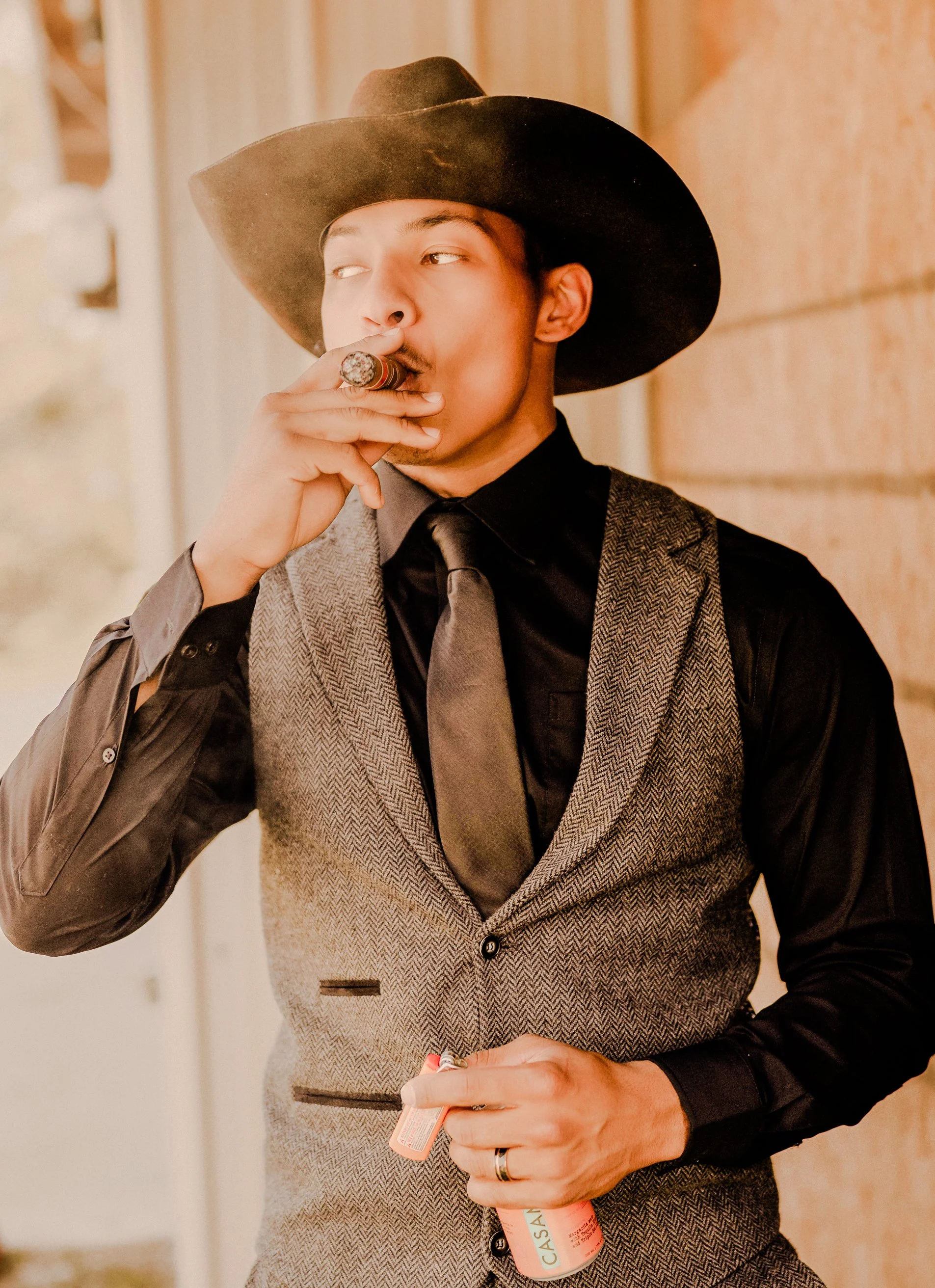 A young man dressed in a black shirt, grey vest, and matching tie, wearing a wide-brimmed black hat, smoking a cigar, holding a bottle in his hand, standing against a brick wall.