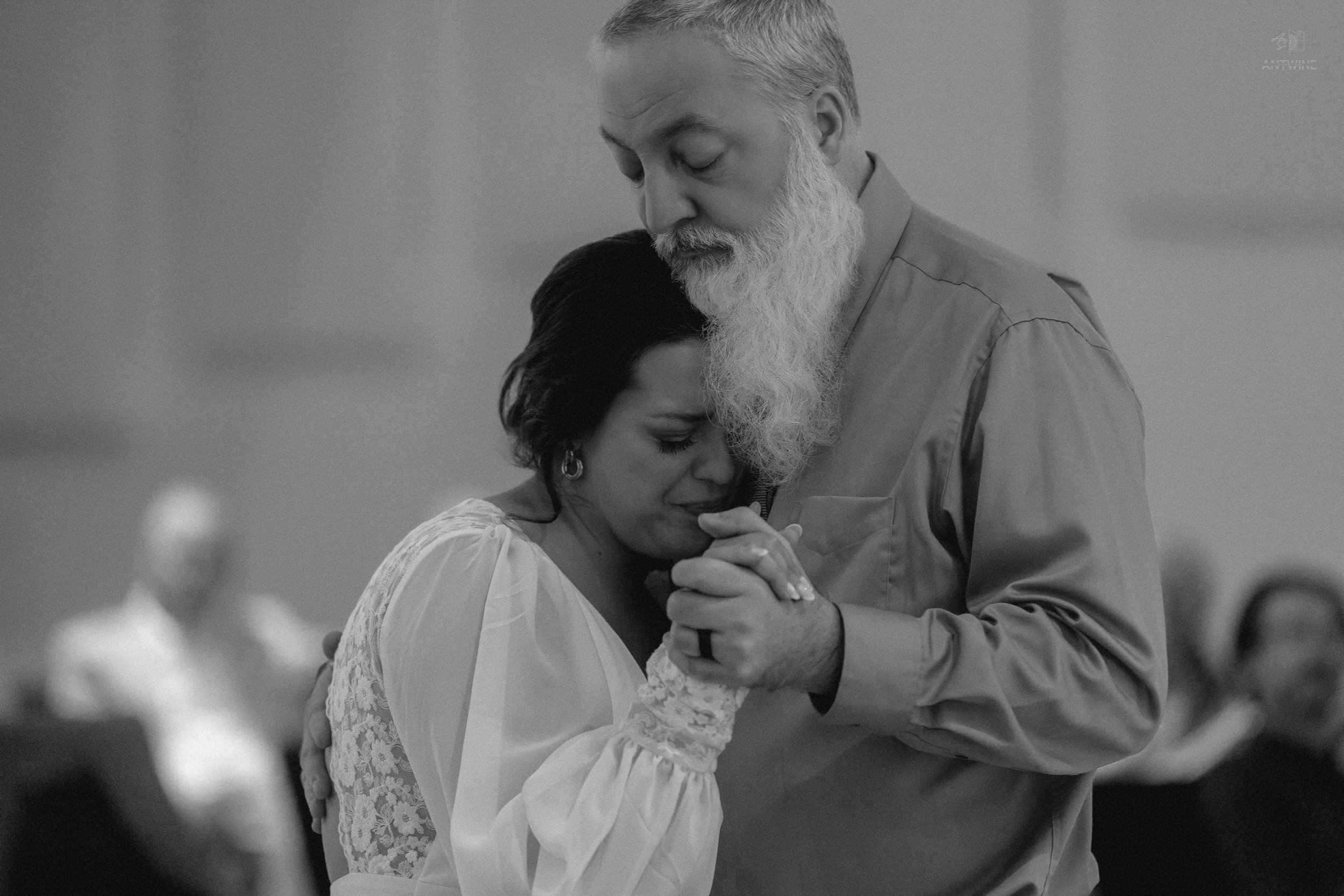 An elderly man with a gray beard dancing with a woman in a white dress. They are holding hands and gently resting their foreheads against each other during a dance.