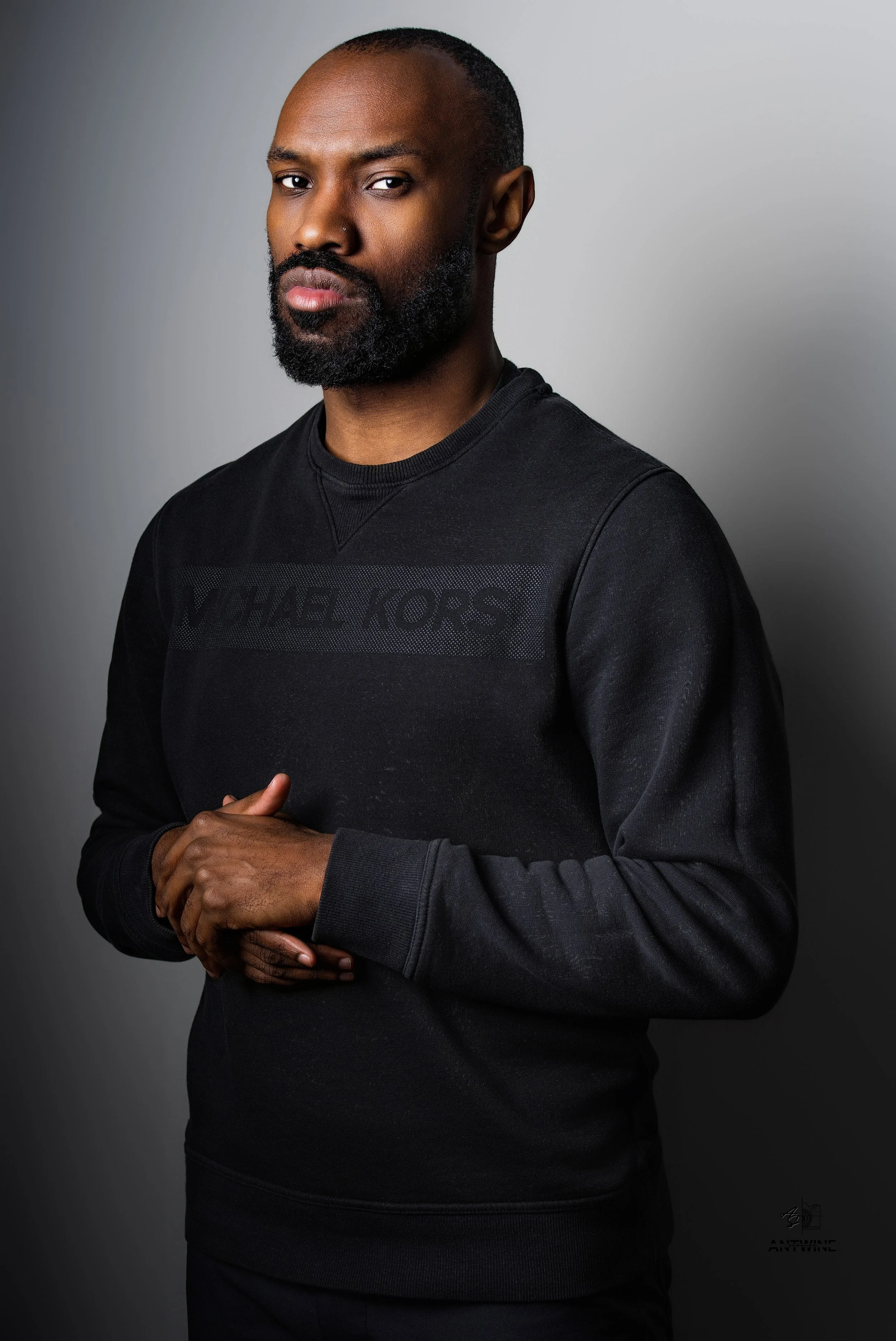A man with a beard and short hair wearing a black Michael Kors sweatshirt, standing against a gray background.