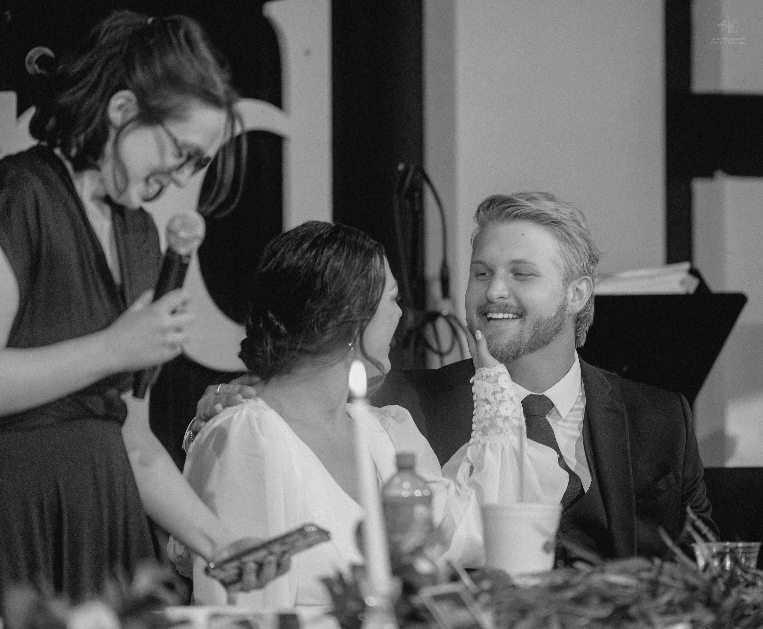 A woman with glasses speaking into a microphone, with her head bent downward. Two other women and a man are seated nearby, engaged and smiling in conversation during an indoor event.