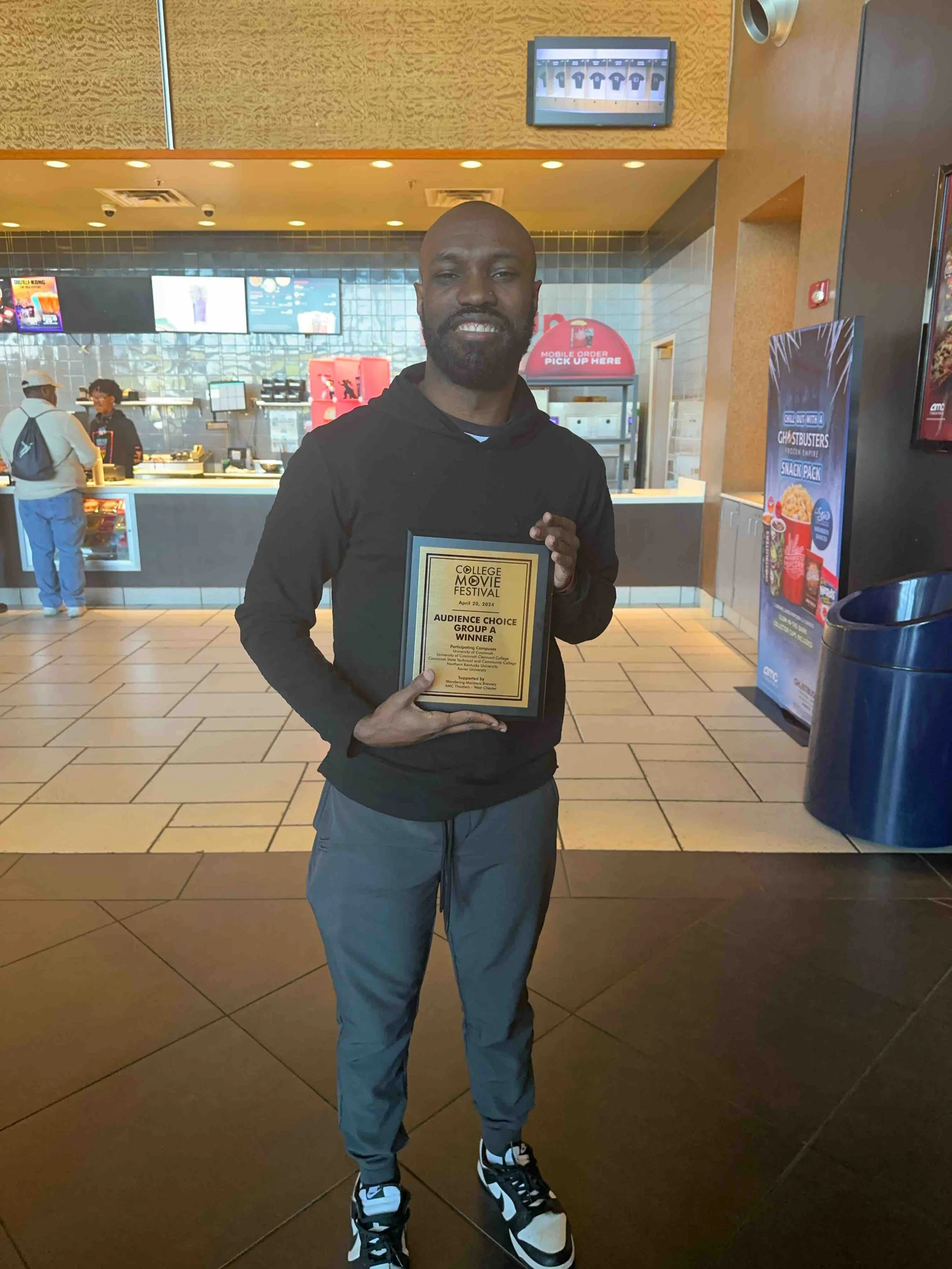 A man standing in a fast-food restaurant holding a plaque that reads 'College Movie Festival, Audience Choice Group A Winner', smiling at the camera.