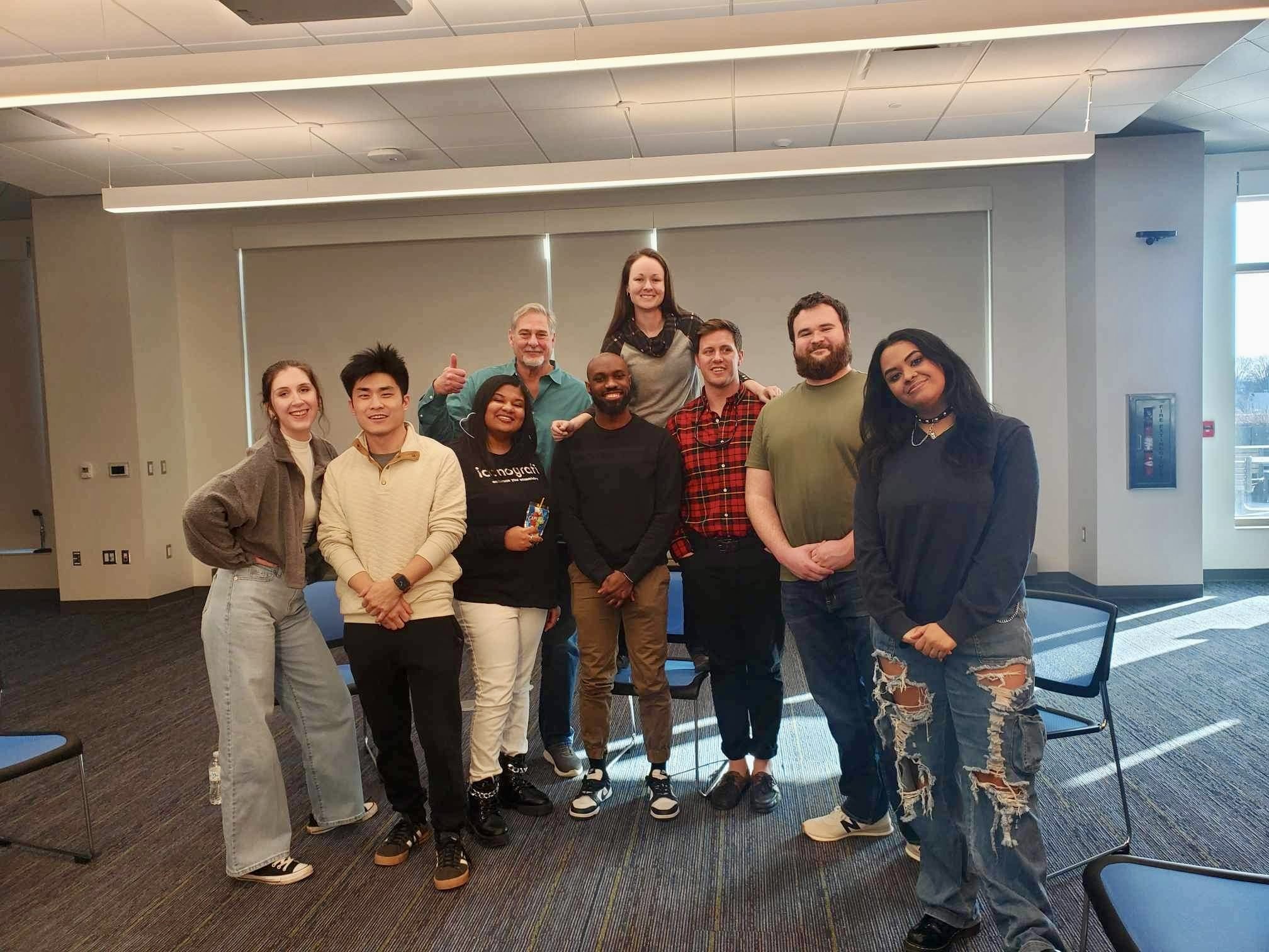 Group photo of ten diverse young adults and one older man smiling and posing together in a modern indoor room with large windows and gray walls.