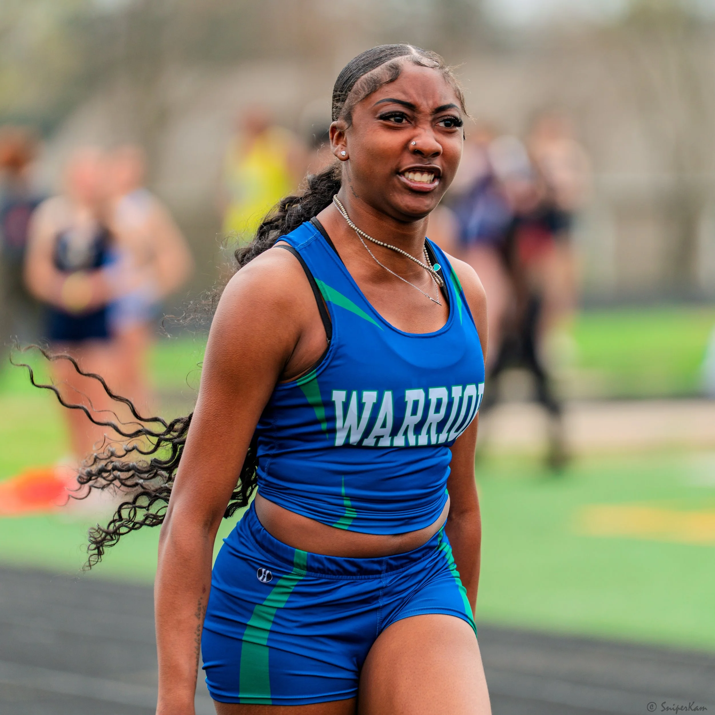 A female sprinter wearing a blue and green athletic uniform with the word 'WARRIORS' on it, running on a track and looking determined.