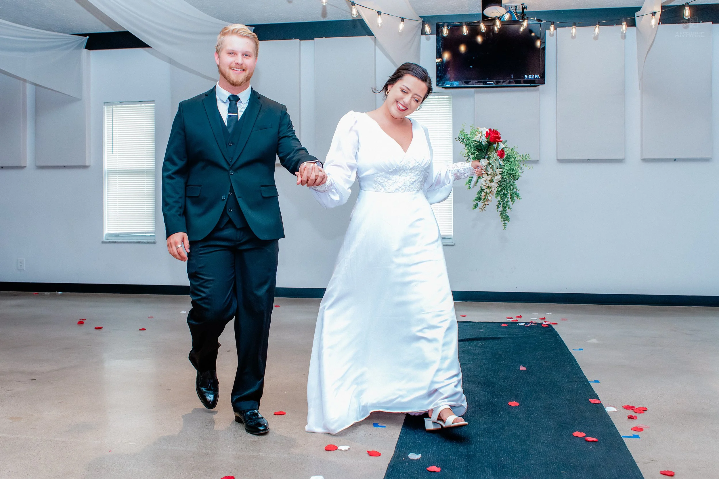 A couple dressed in wedding attire holding hands and walking down an aisle decorated with rose petals, with the groom smiling and the bride holding a bouquet, in an indoor wedding ceremony.