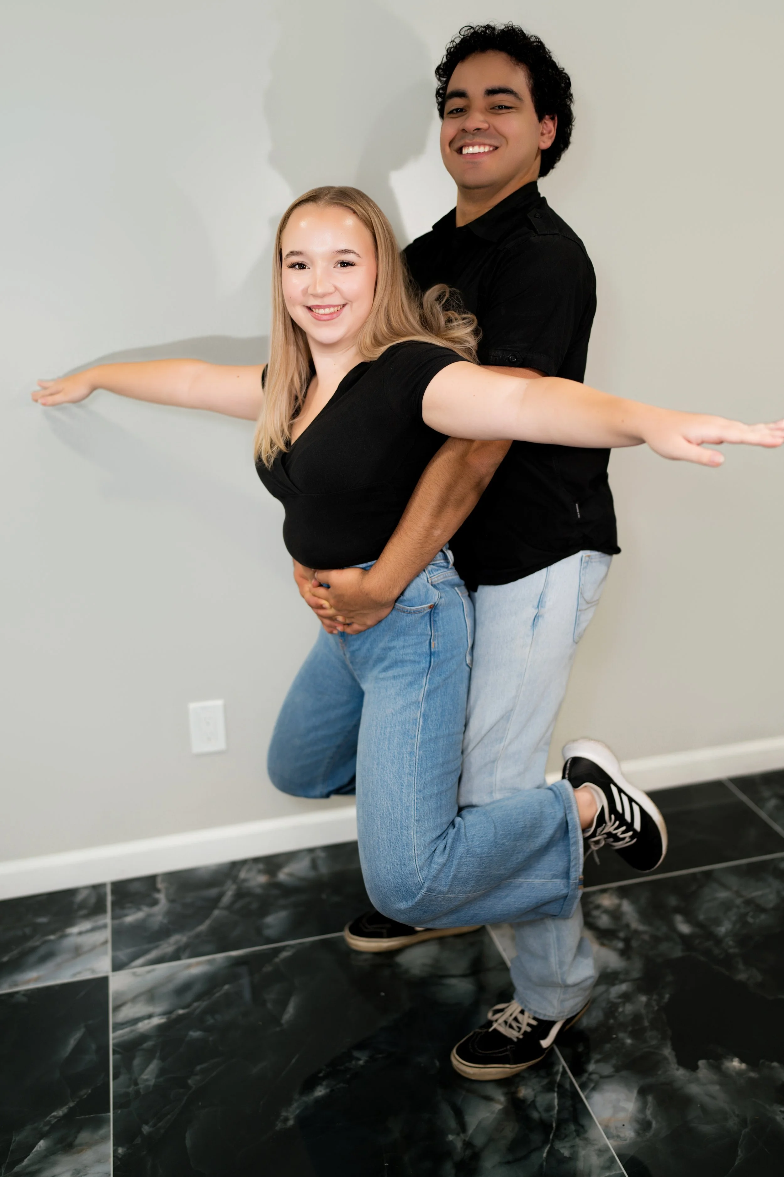 A young woman and a young man are smiling and posing playfully against a plain wall, with the man holding the woman around her waist and her arm extended outward as if balancing.