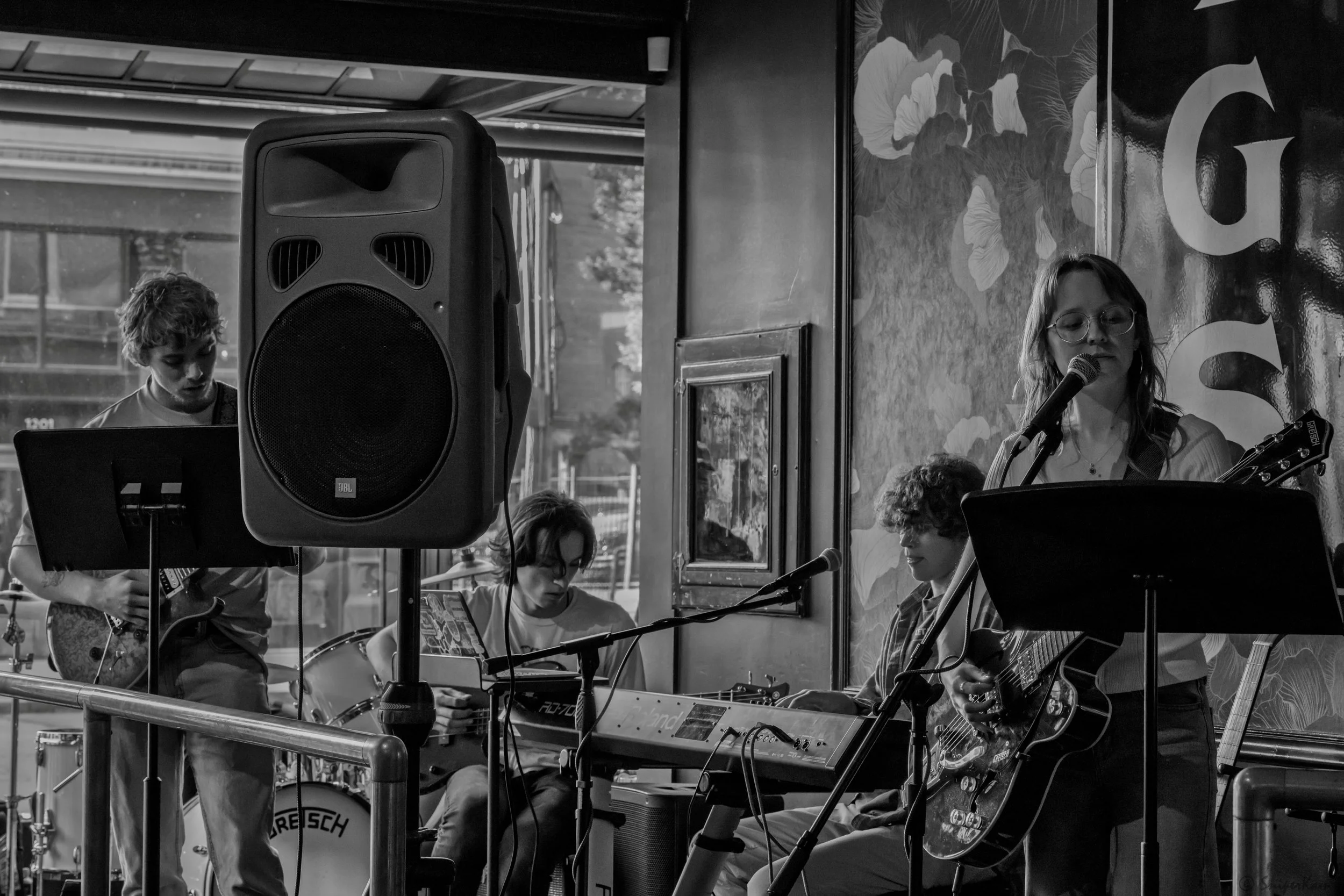 A band performing live indoors, featuring a female singer with glasses and long hair playing guitar, a keyboardist, a bassist, and a drummer, with a large speaker in the foreground.
