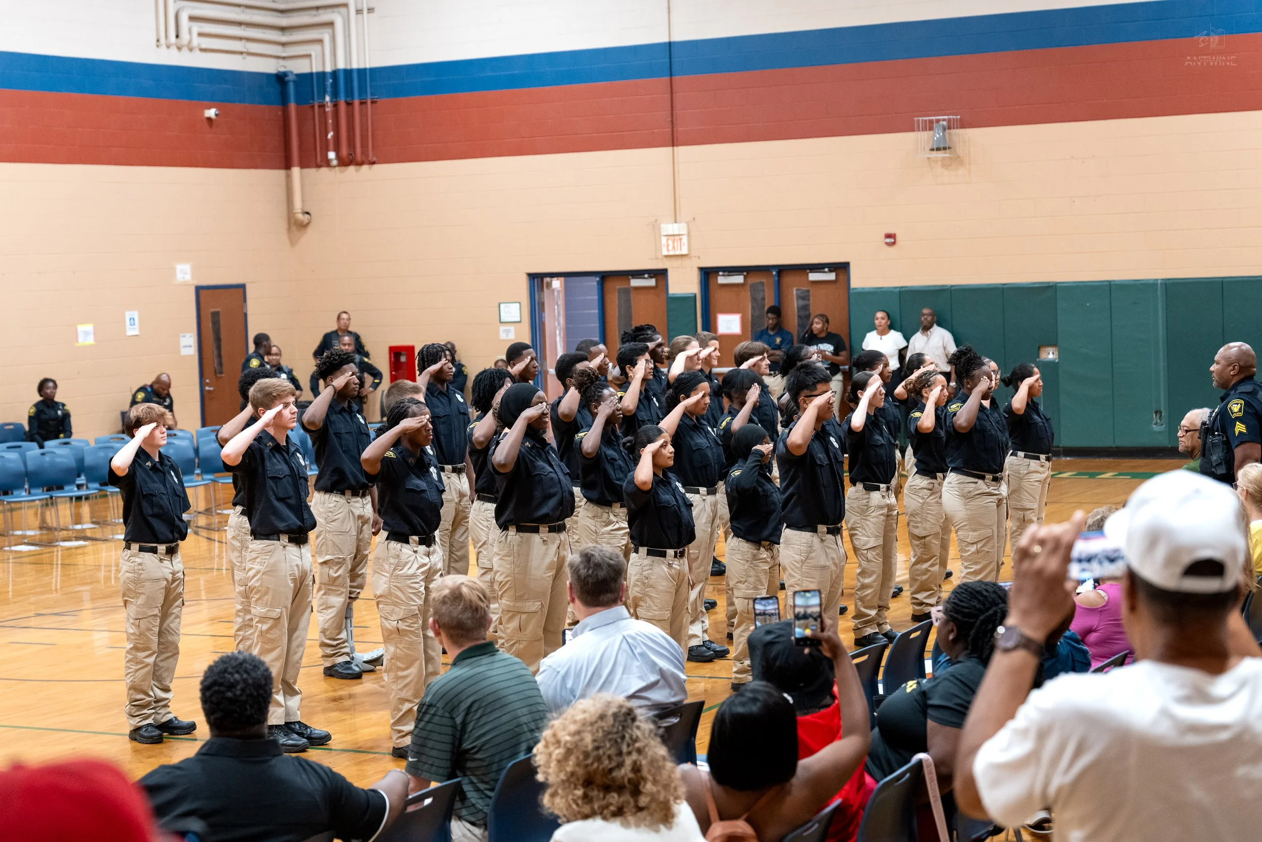 Group of young people in uniform standing and saluting inside a gymnasium during a ceremony or event, with seated audience watching.