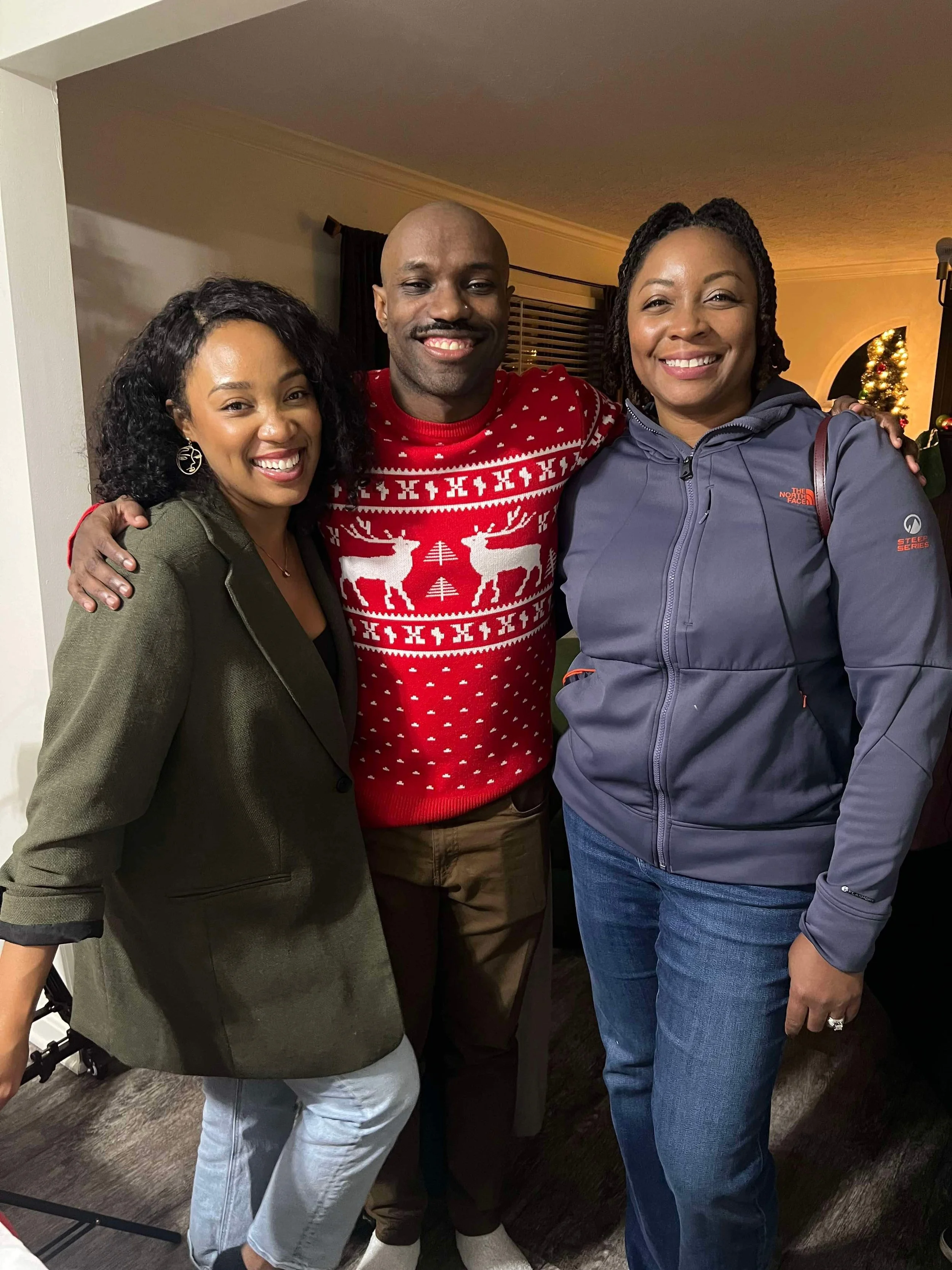 Three people smiling and hugging each other indoors, with a Christmas tree in the background, celebrating the holiday season.