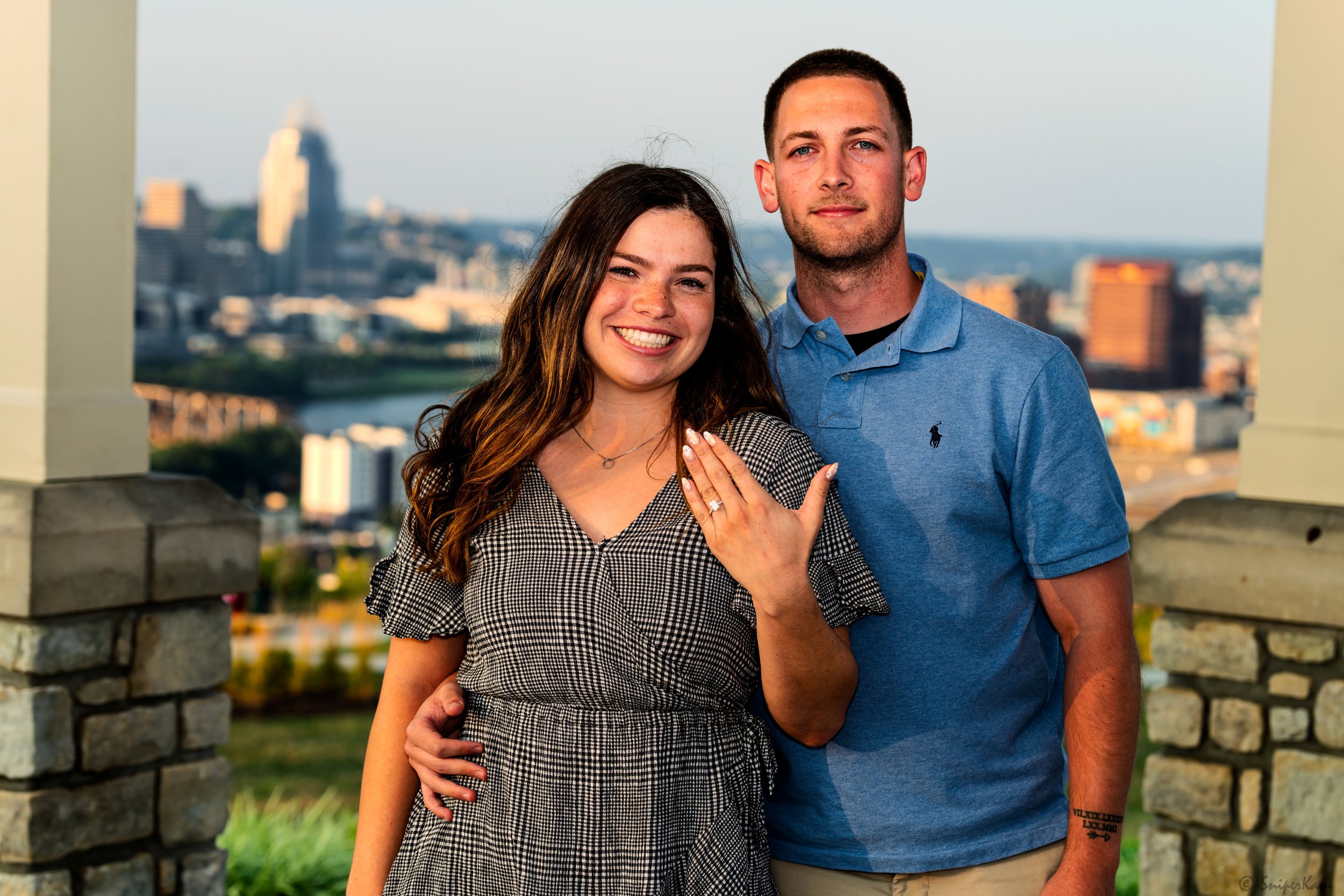 A smiling woman showing off an engagement ring on her left hand, standing next to a man with an arm around her, on a balcony overlooking a city skyline at sunset.