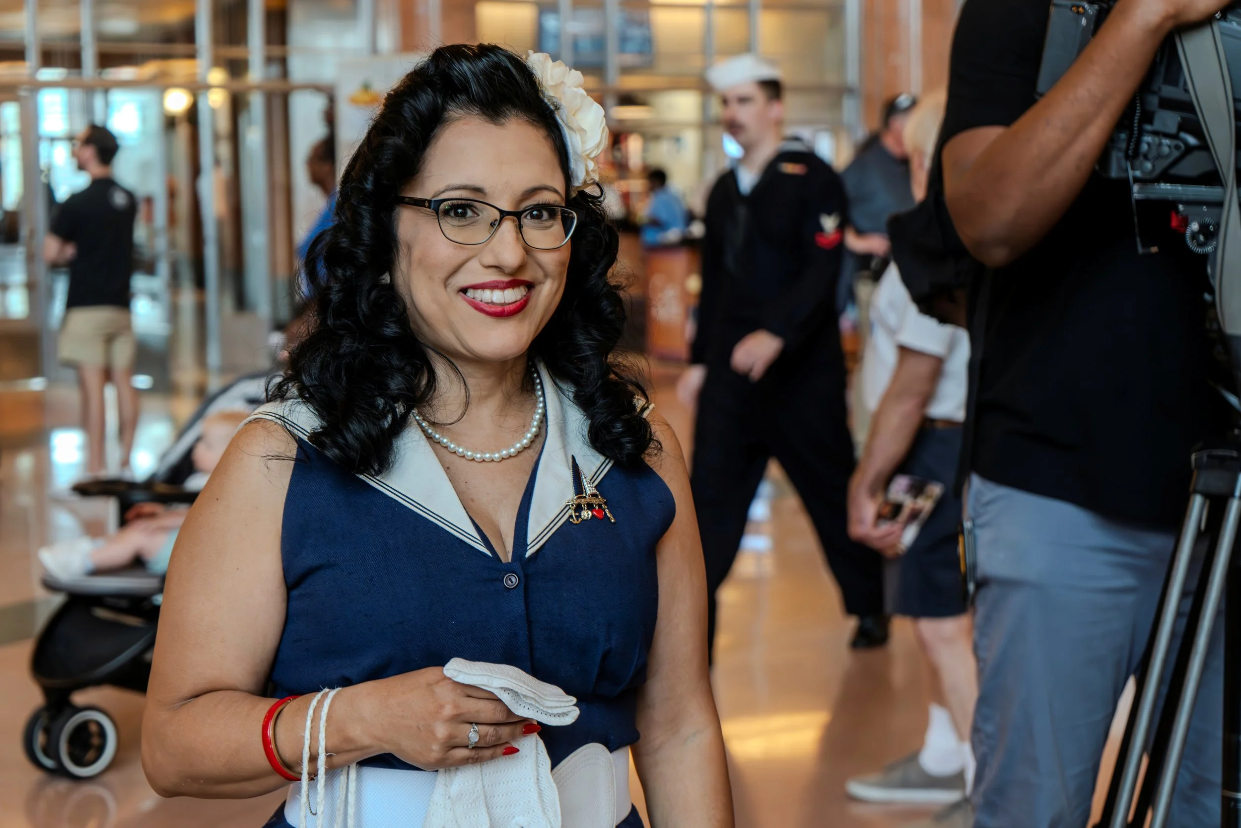 A woman smiling at a camera, dressed in vintage-style clothing with a pearl necklace, glasses, and a white flower in her curly dark hair, standing indoors with a busy background of people and a stroller.