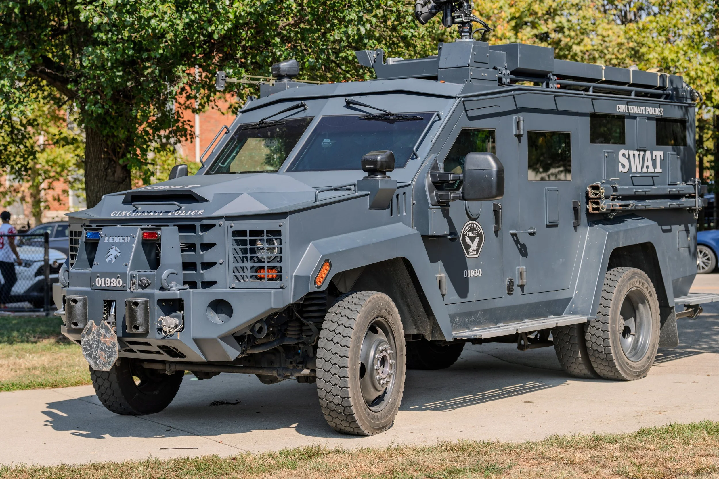 A gray SWAT armored police vehicle parked on a sidewalk, with trees and a fence in the background.