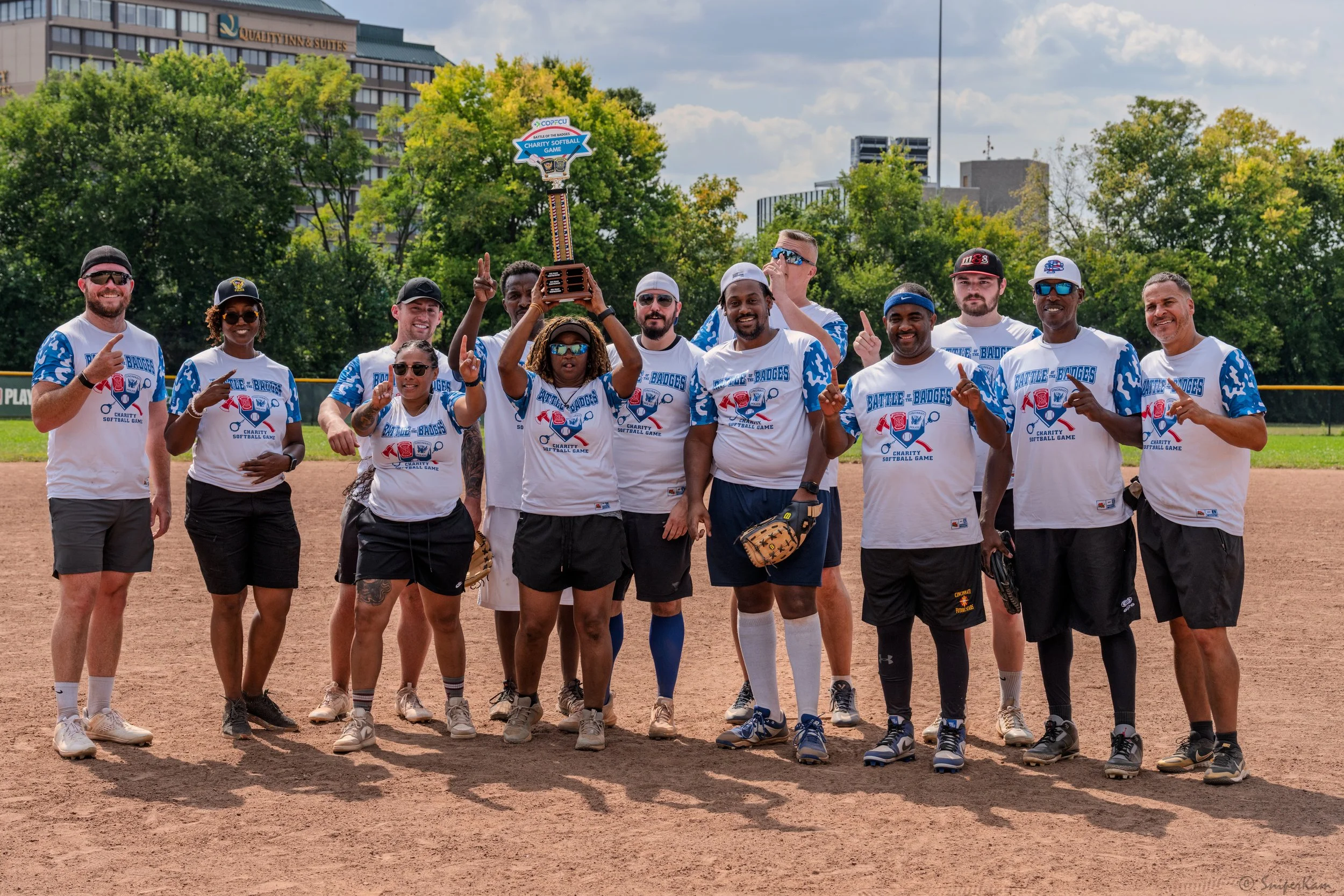 A group of people standing on a baseball field, holding a trophy, celebrating after a softball game. They are wearing matching white and blue team shirts that say "Battle Badges" and are posing with their hands raised or pointing. Some are wearing su
