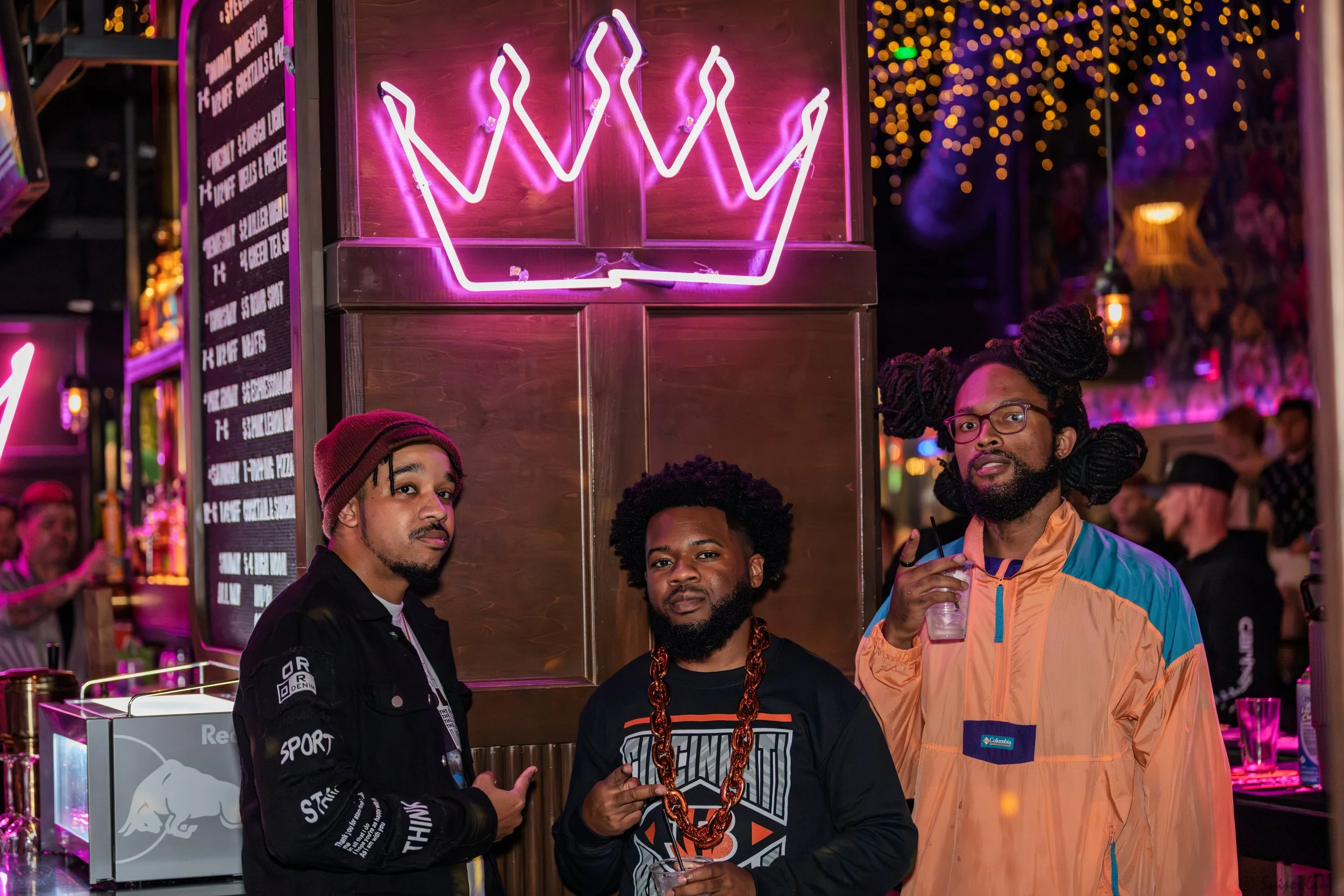 Three men posing at a bar with neon crown sign above them in a lively, colorful nightlife setting.