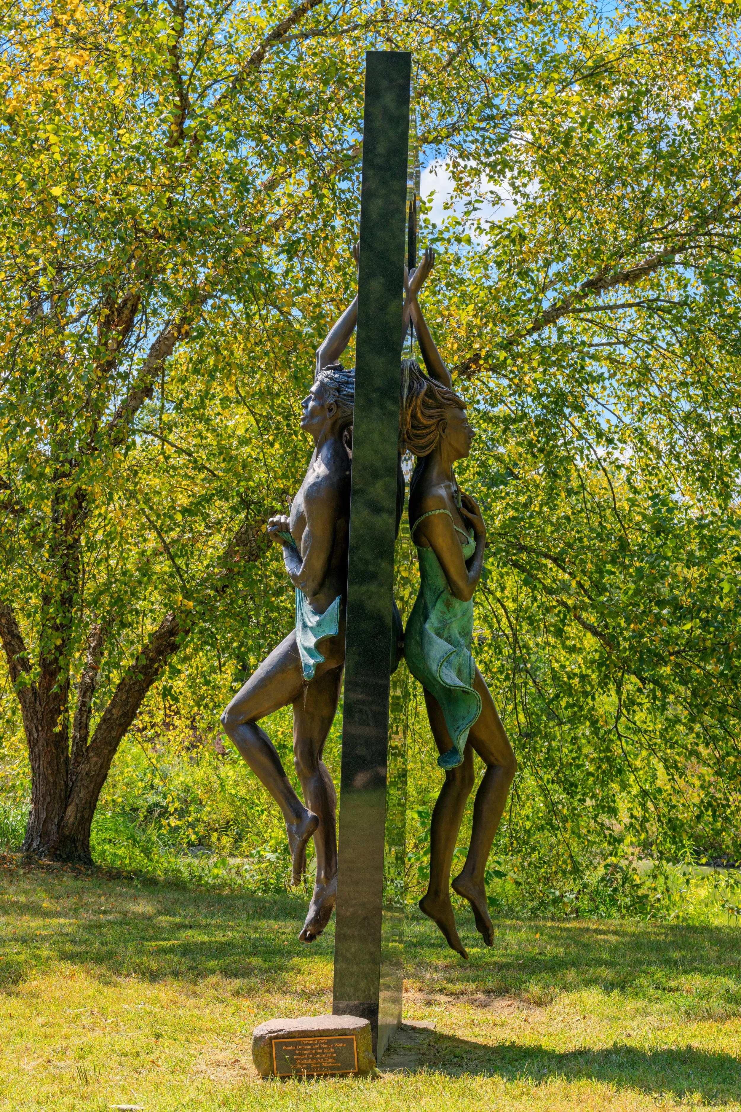 Sculpture of two women hanging upside down, back to back, with a vertical reflective panel between them, situated outdoors with trees and grass in the background.