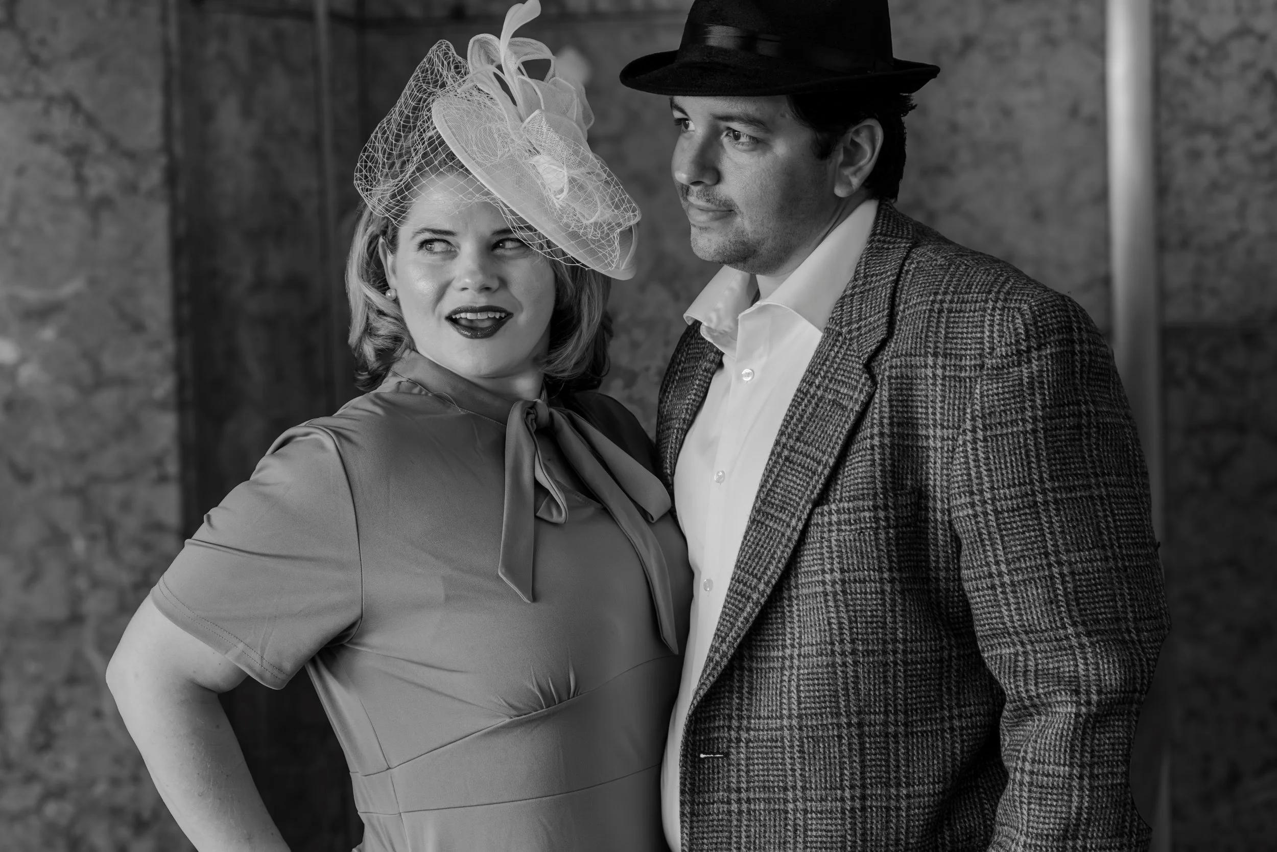 A woman in a vintage dress with a headscarf and veil, and a man in a suit and hat, standing together against a brick wall.