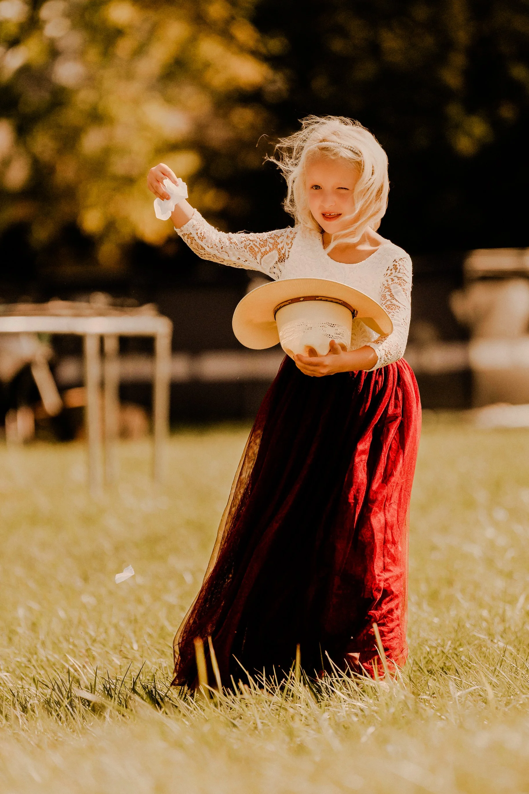 A young girl with blonde hair in a white lace top and red velvet skirt playing with a pie in an outdoor setting.