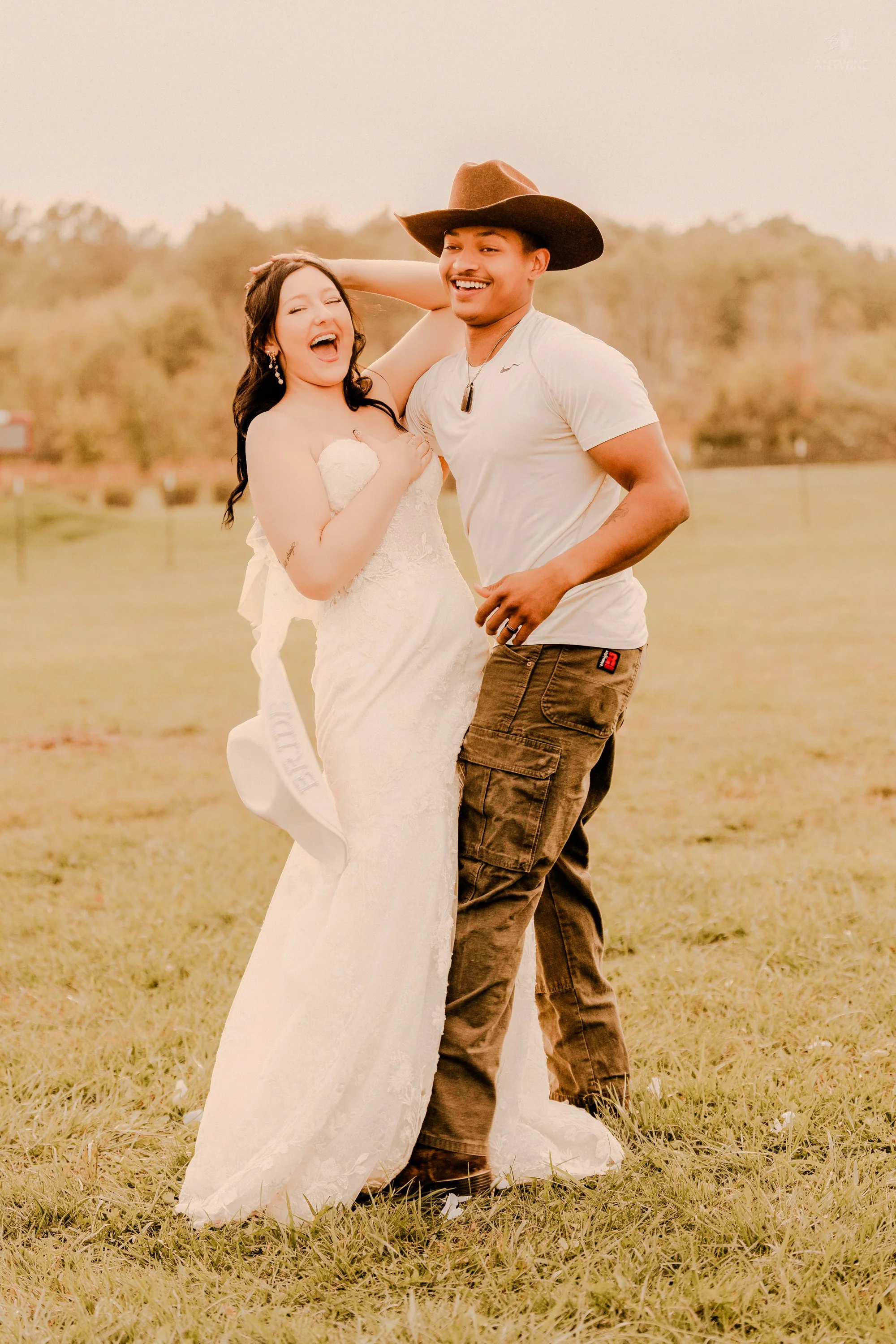 A happy couple outdoors, with the woman wearing a white wedding dress and the man in casual attire and a cowboy hat.