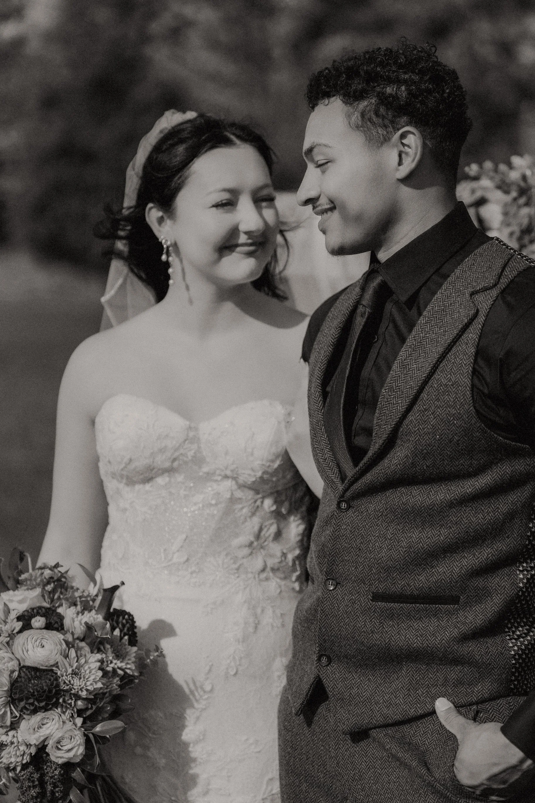 A black and white photo of a smiling bride in a strapless lace wedding dress with a bouquet, standing next to a groom in a dark shirt and patterned vest, outdoors.