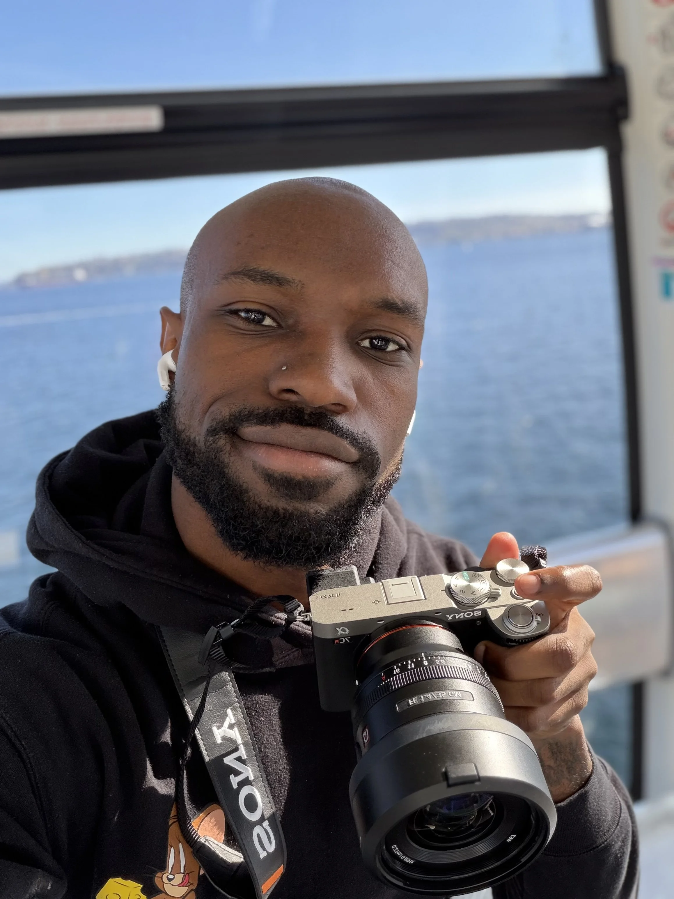 A man with a bald head, beard, and nose piercing taking a selfie with a camera on a boat with water and distant land in the background.