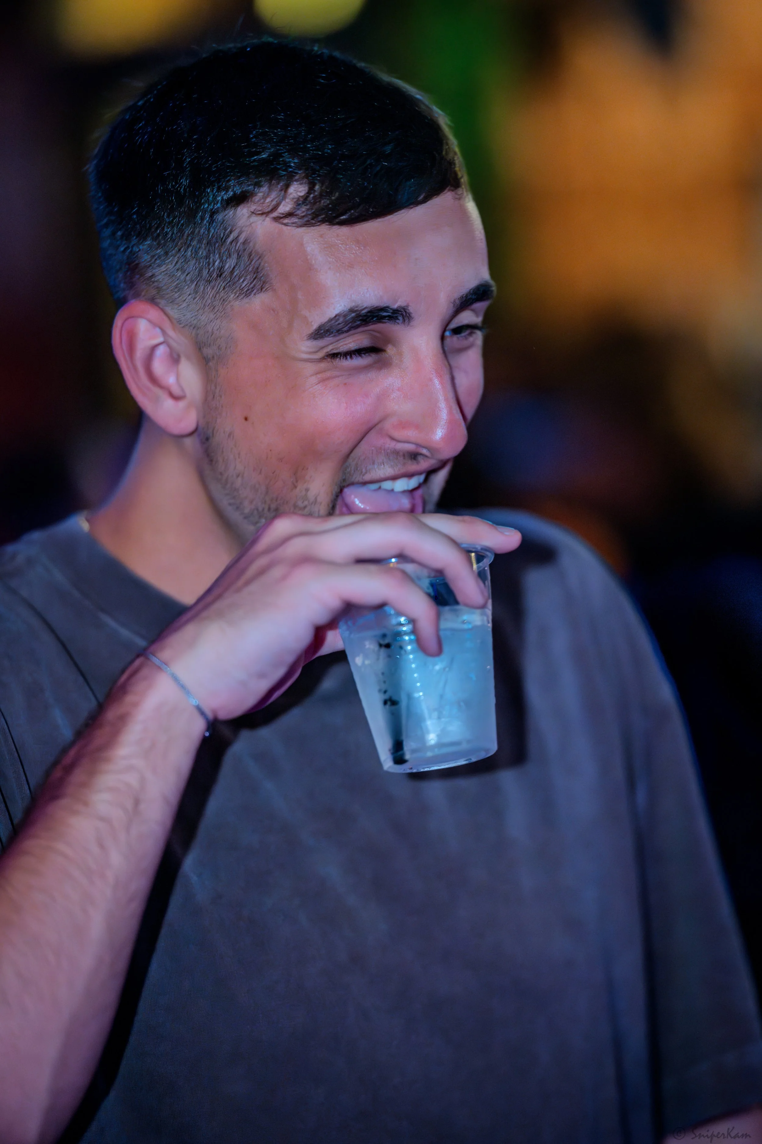 A young man with short dark hair and a slight beard smiling and winking at the camera while holding a glass of clear beverage with ice.