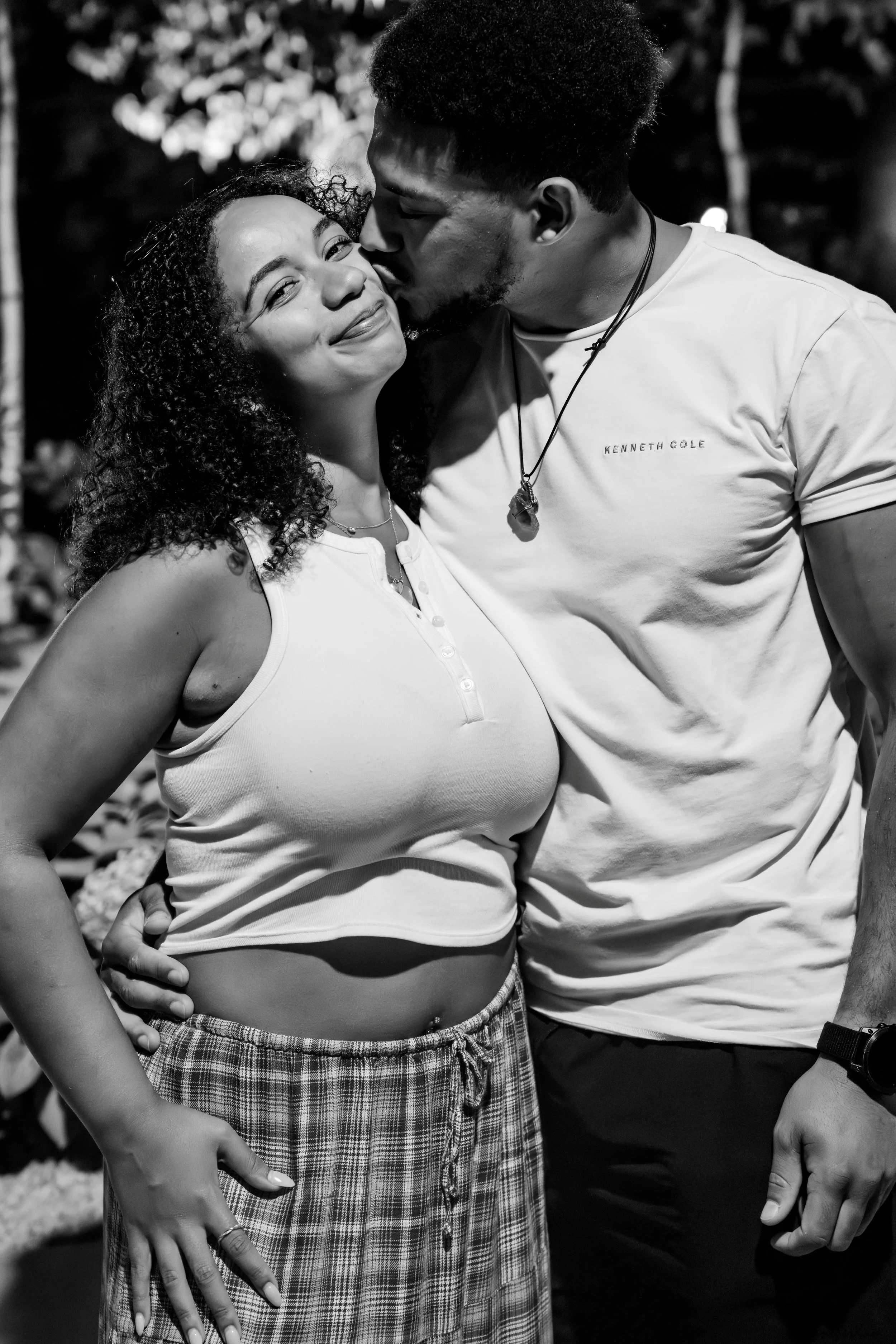 A black and white photo of a man kissing a woman on the cheek. The woman is smiling with closed eyes, wearing a sleeveless top and plaid pants, with her hand on her hip. The man is wearing a T-shirt, has face tattoos, and is wearing a necklace. They 