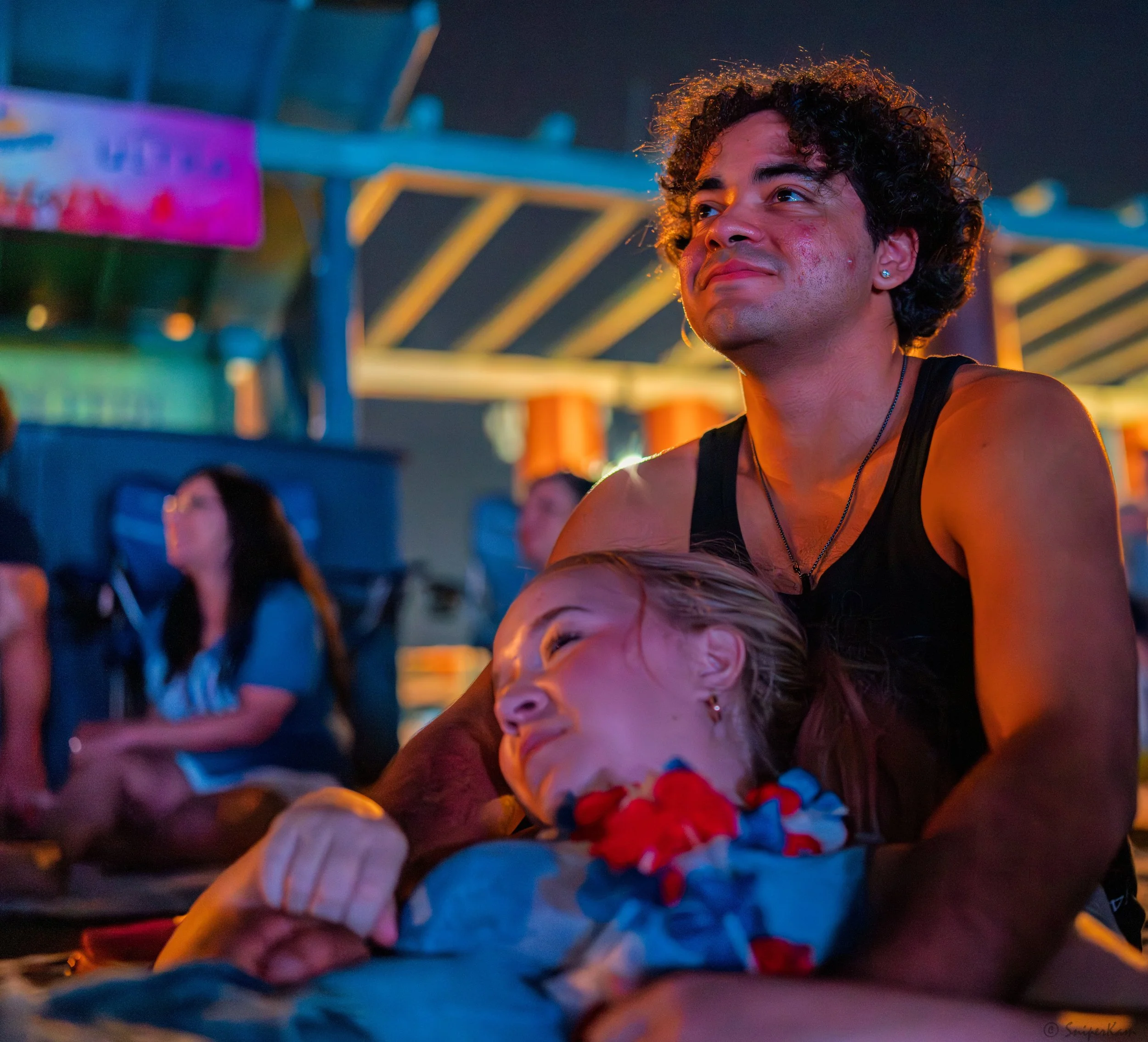 A man and a woman sharing a tender moment at a carnival or fair, with the woman resting her head on the man's lap and looking up at him, while the man looks contemplative. The background has colorful lights and other people sitting and relaxing.