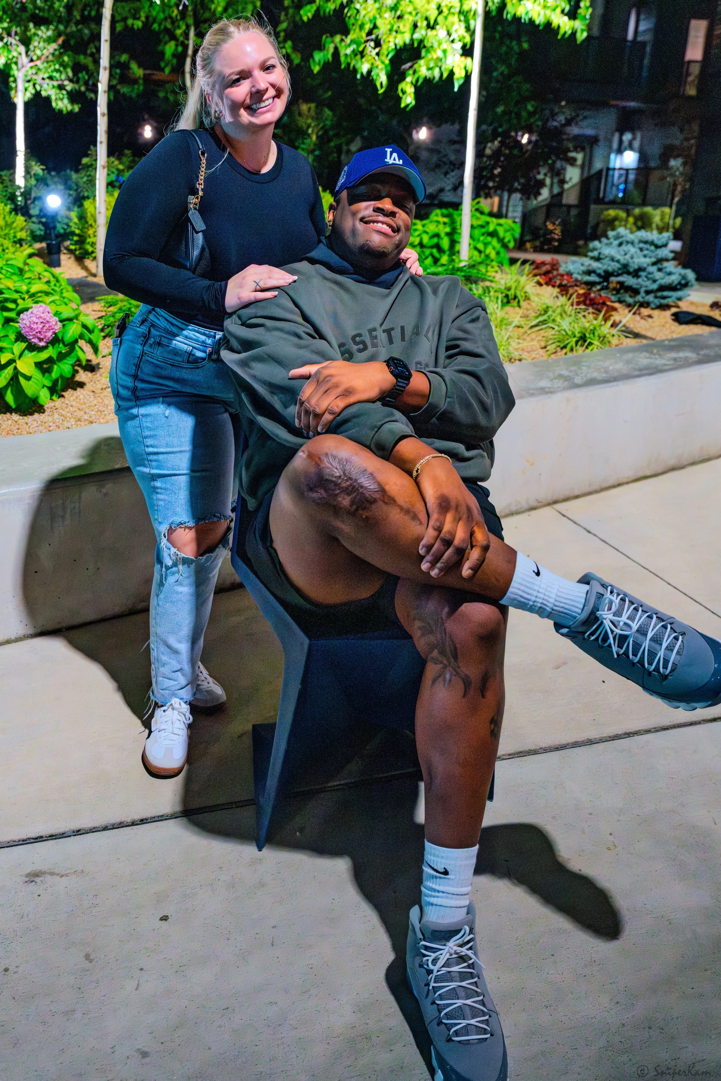 A smiling woman with blonde hair standing next to a seated man with a knee injury, outdoors at night in front of greenery.