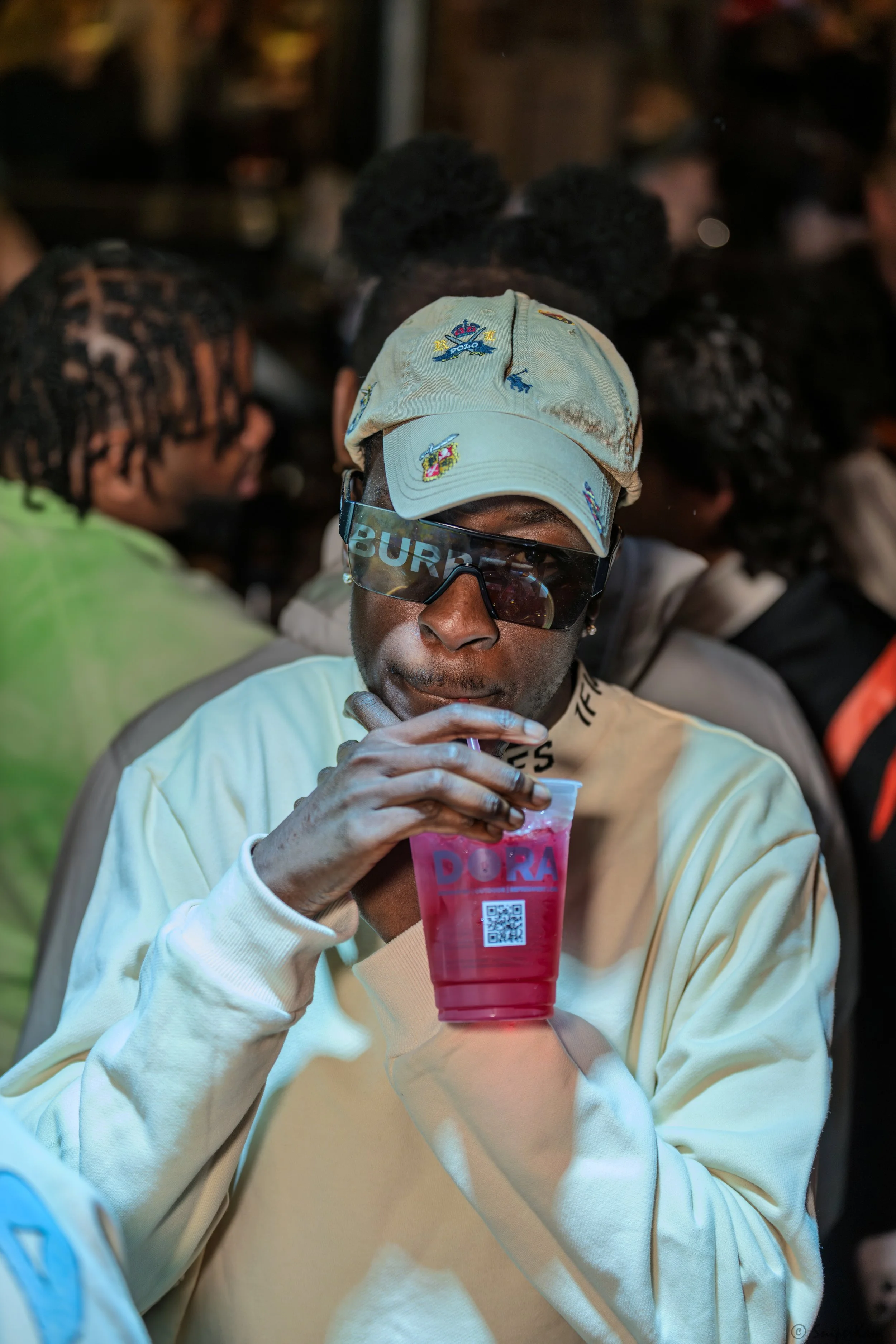 A person wearing a beige baseball cap, dark sunglasses, and a white sweatshirt, holding a pink drink with the label 'DORA' in a social setting.