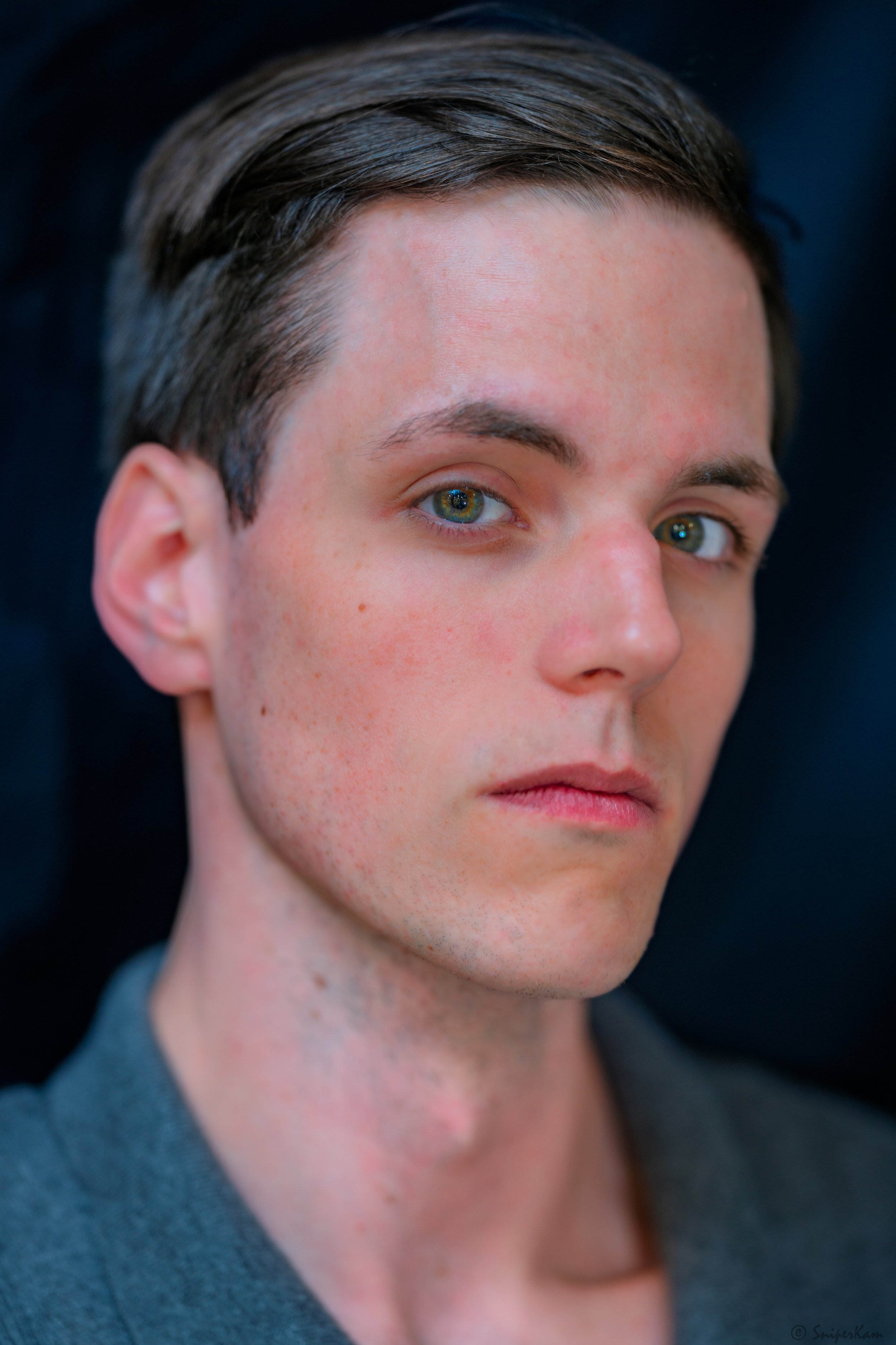 Close-up portrait of a young man with light skin, dark brown hair, and blue eyes, looking directly at the camera against a dark background.