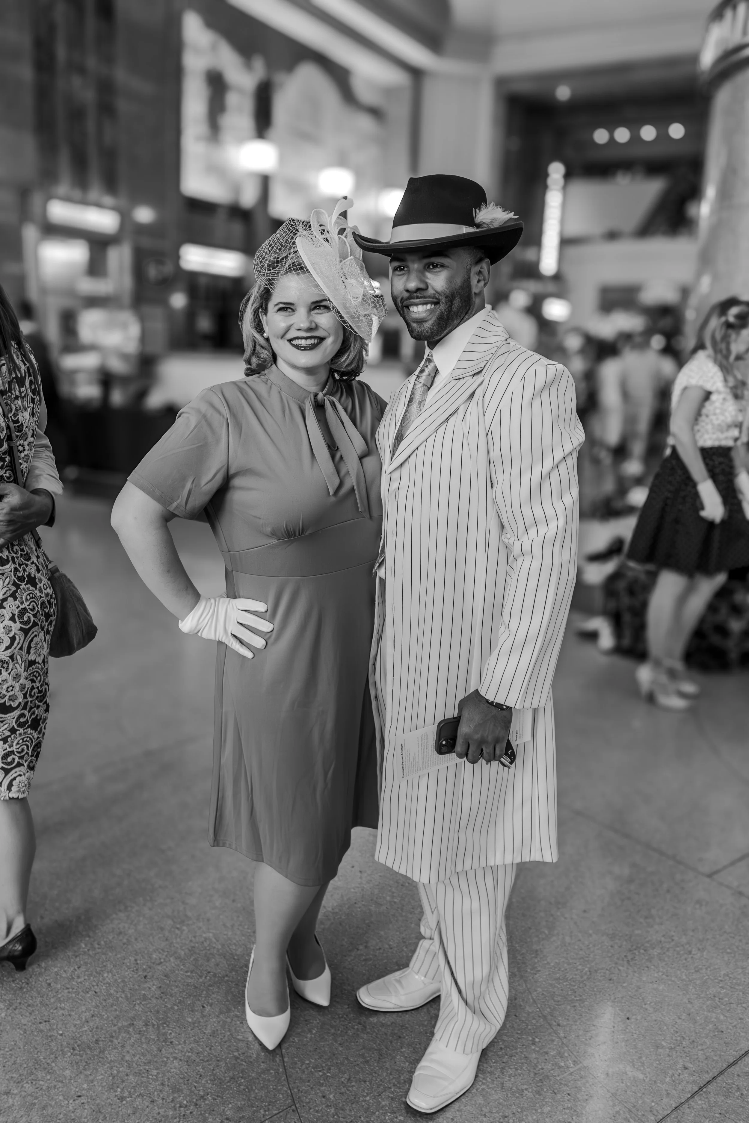A woman and a man dressed in vintage-style clothing, standing together and smiling in a crowded indoor setting.