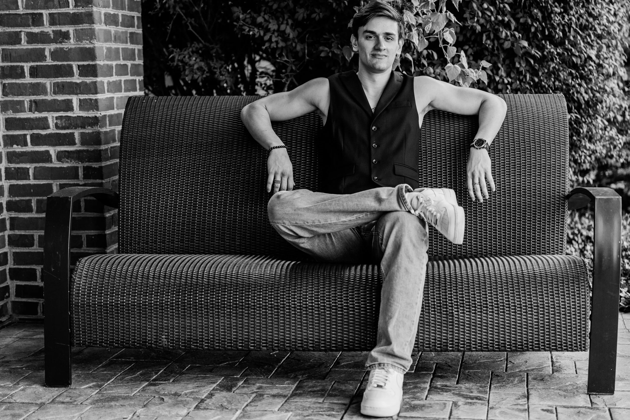 A young man with short hair sitting on a park bench, wearing a sleeveless vest, jeans, and sneakers, with one arm resting on the back of the bench and the other on his knee, outdoors with a brick wall and leafy background.