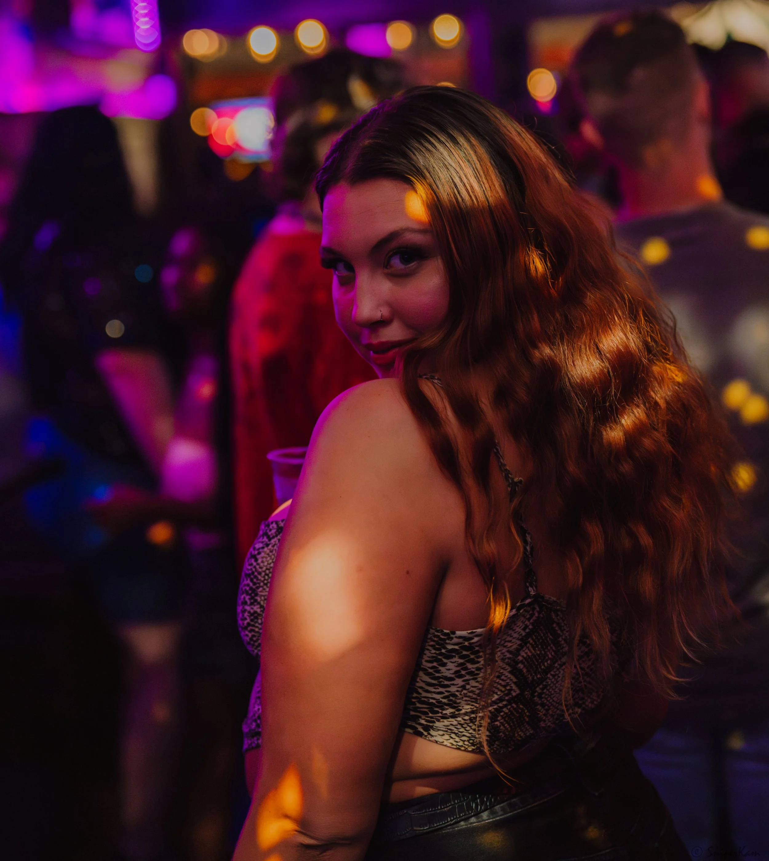 A woman with long wavy red hair looking over her shoulder at a lively party or club scene with colorful lights and other people in the background.