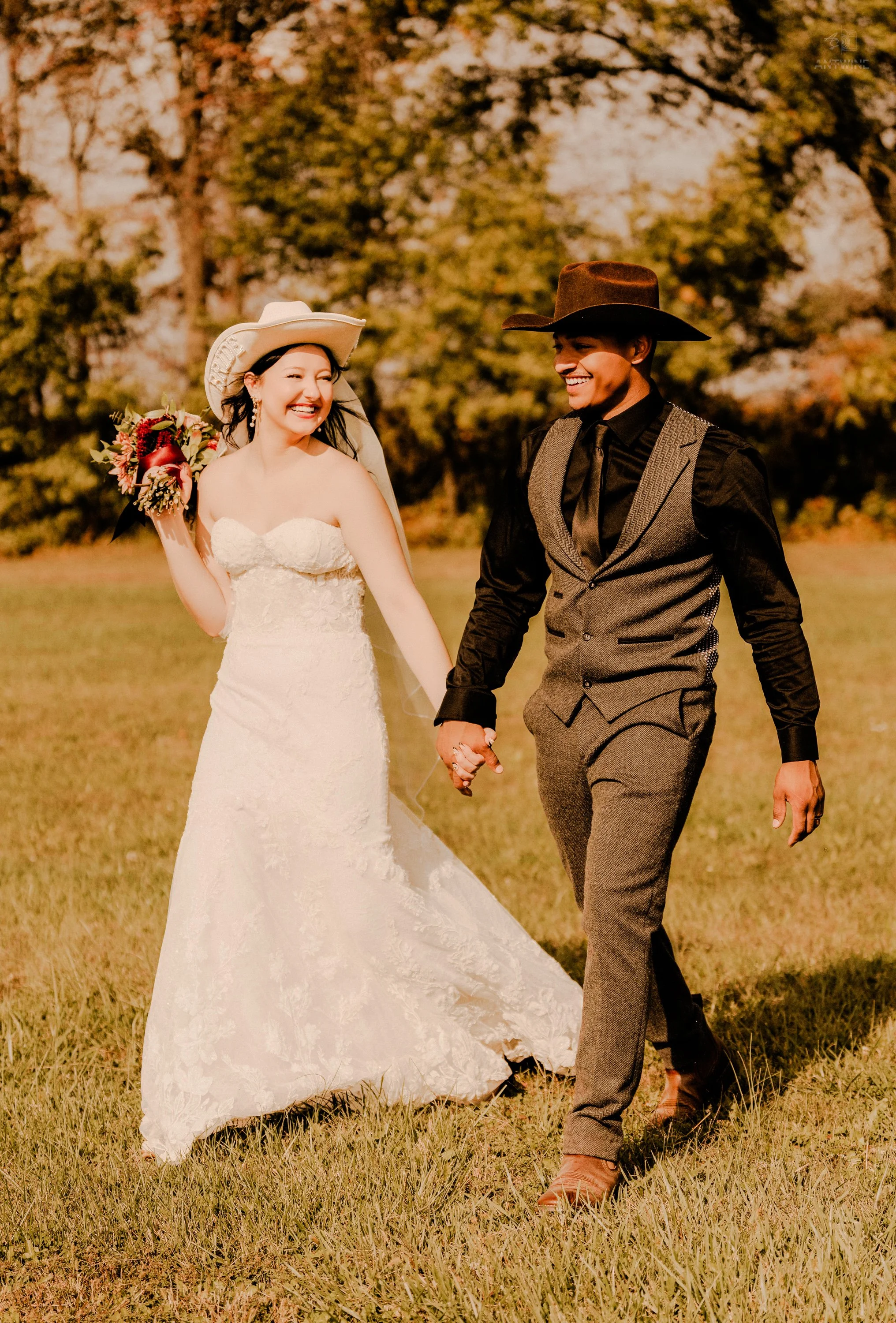 A bride in a white wedding dress holding a bouquet and a groom in a dark shirt and gray vest holding hands, walking outdoors on green grass with trees in the background, during sunset.