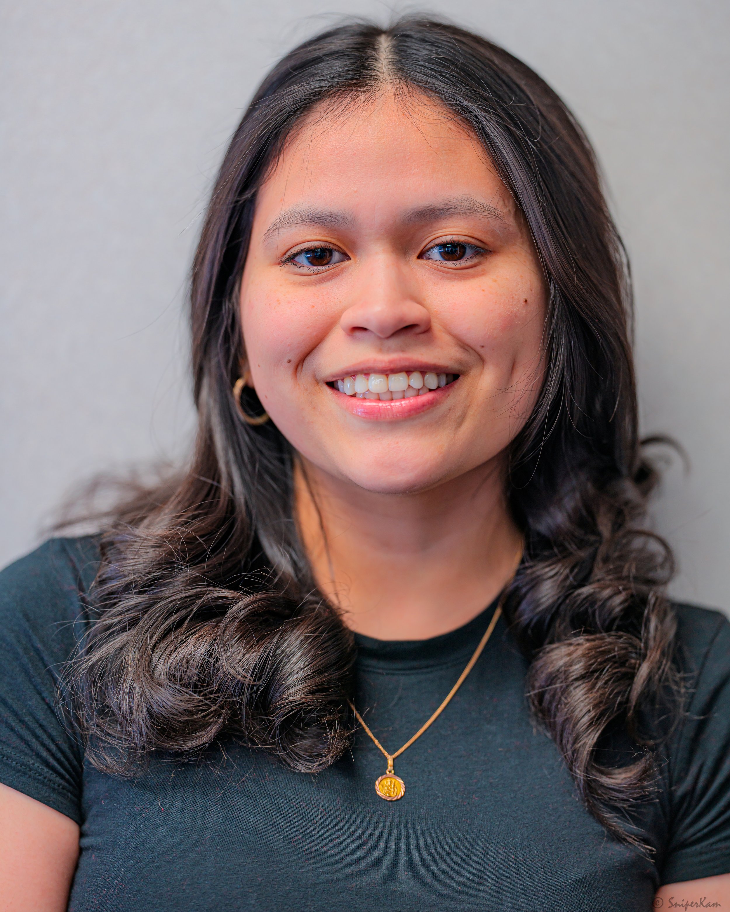 A smiling woman with wavy dark hair, wearing a black shirt, gold hoop earrings, and a gold necklace.
