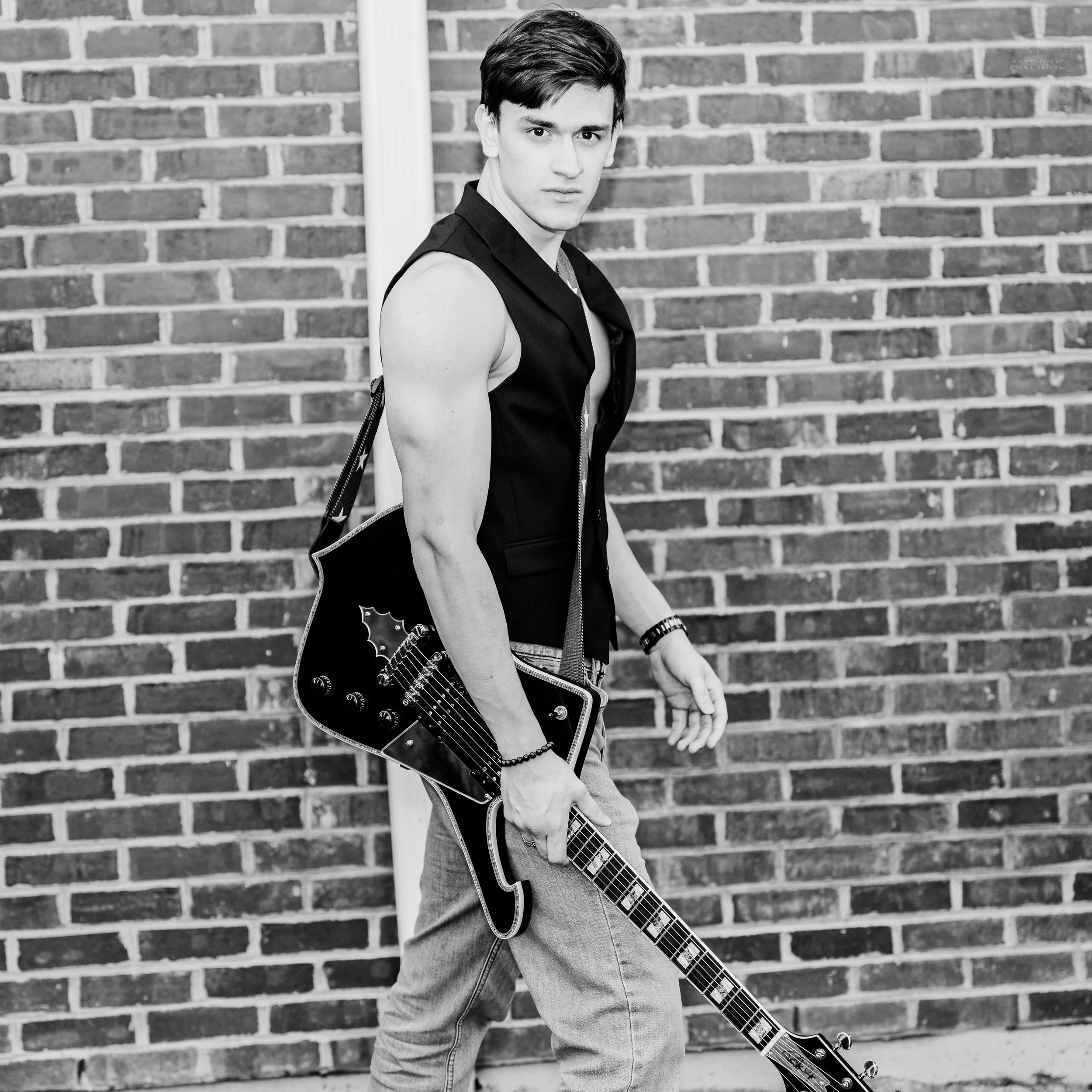 Black and white photo of a young man with short dark hair, wearing a sleeveless vest and jeans, holding an electric guitar, standing against a brick wall.