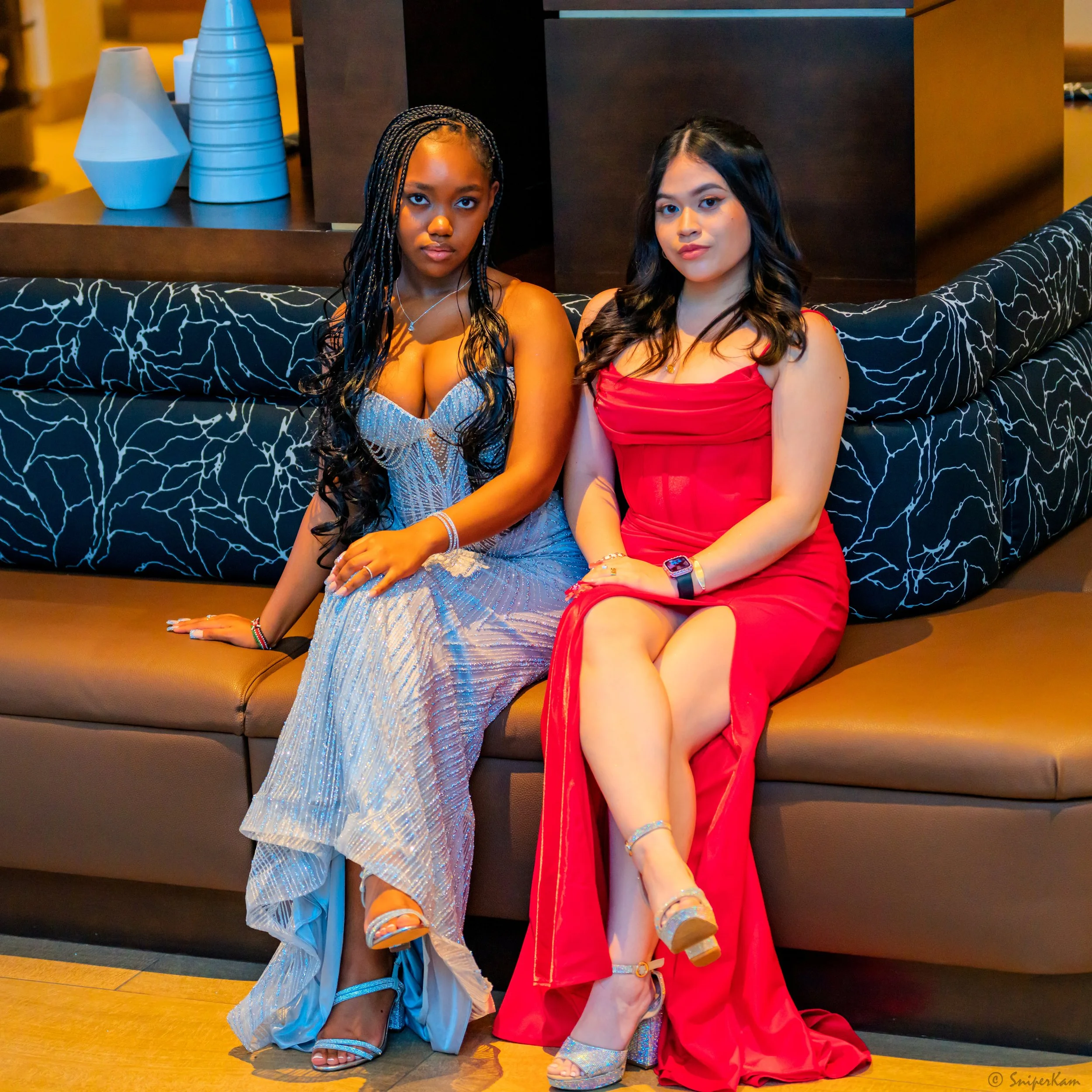 Two women sitting on a brown couch in a stylish indoor setting. The woman on the left is wearing a silver, strapless dress with a slit and has long, curly black hair. The woman on the right is wearing a red dress with a slit and has shoulder-length b