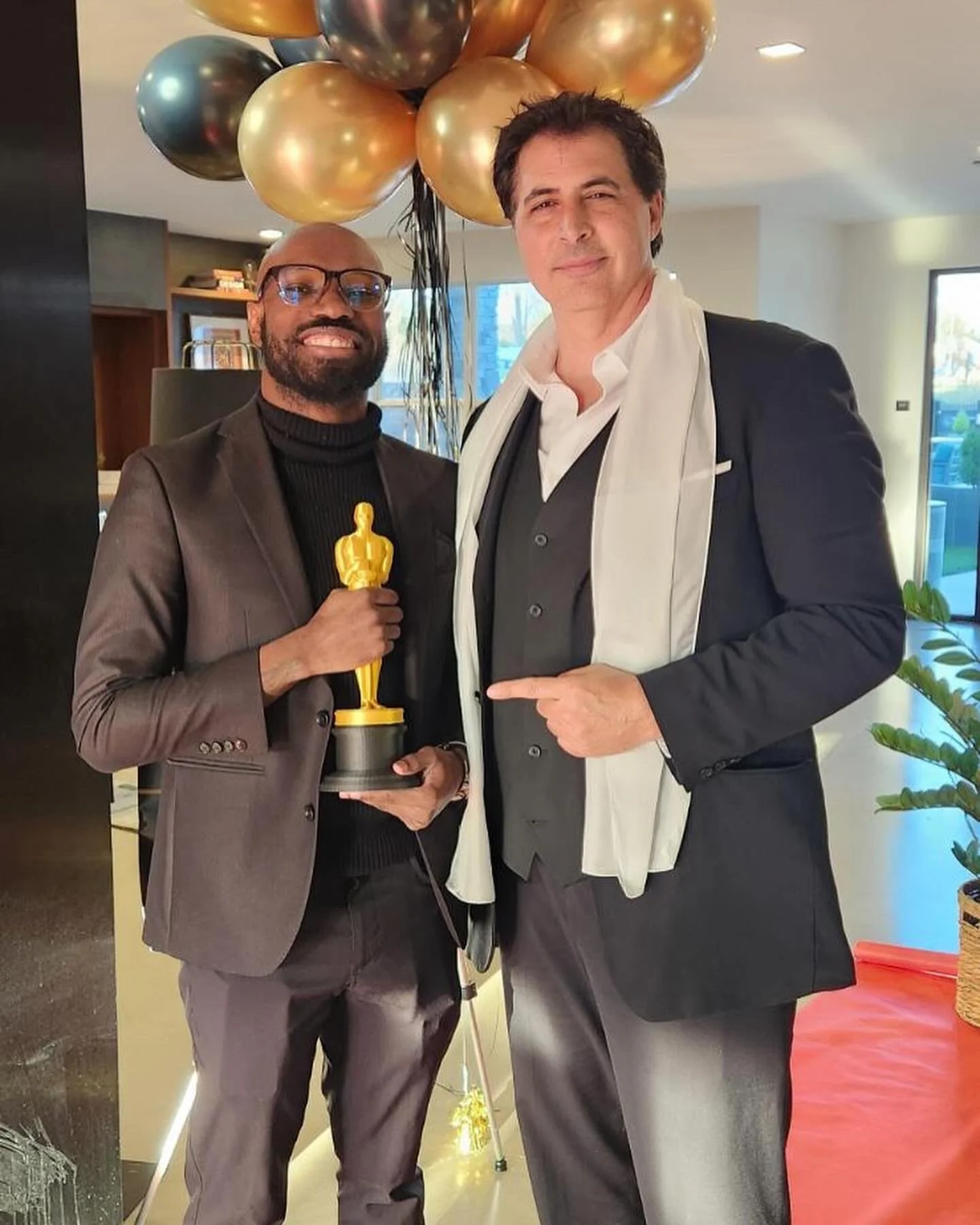 Two men in suits standing indoors, one holding a small Oscar-like trophy, with balloons in gold, black, and bronze behind them.